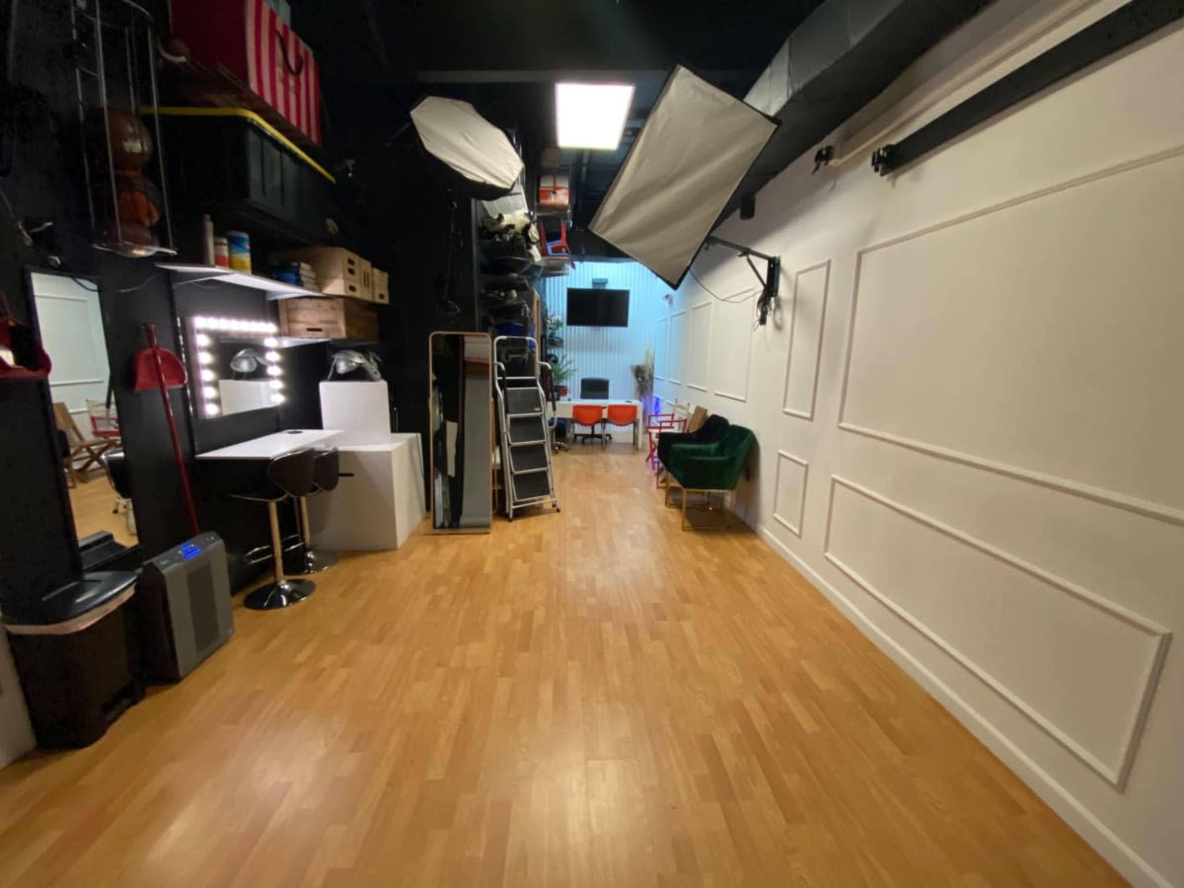 A studio space with wooden flooring, studio lighting equipment, and a mix of furniture including a bar stool, a ladder, and a green velvet chair.