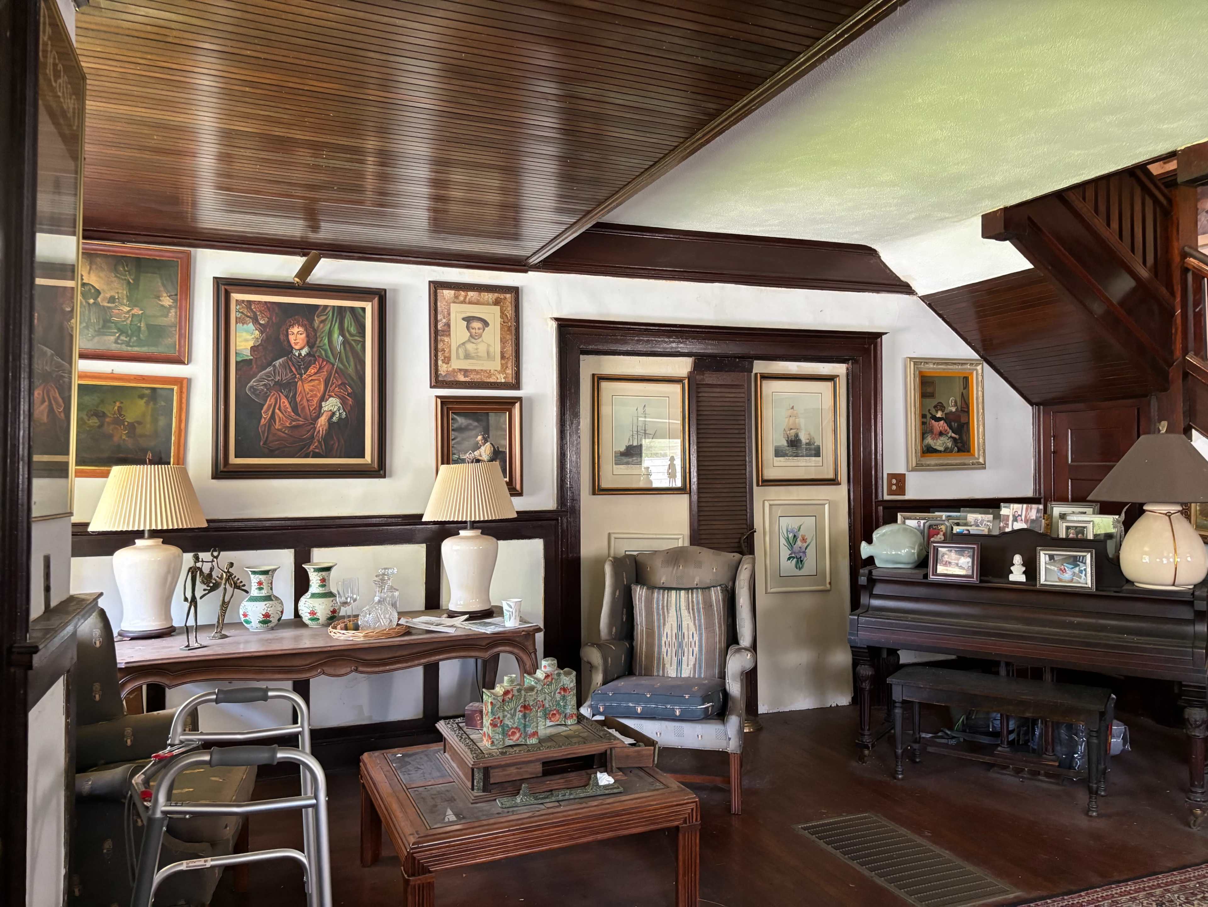 The image shows a vintage interior with framed paintings on the walls, a piano, and a mix of antique furniture, including a table with decorative items.