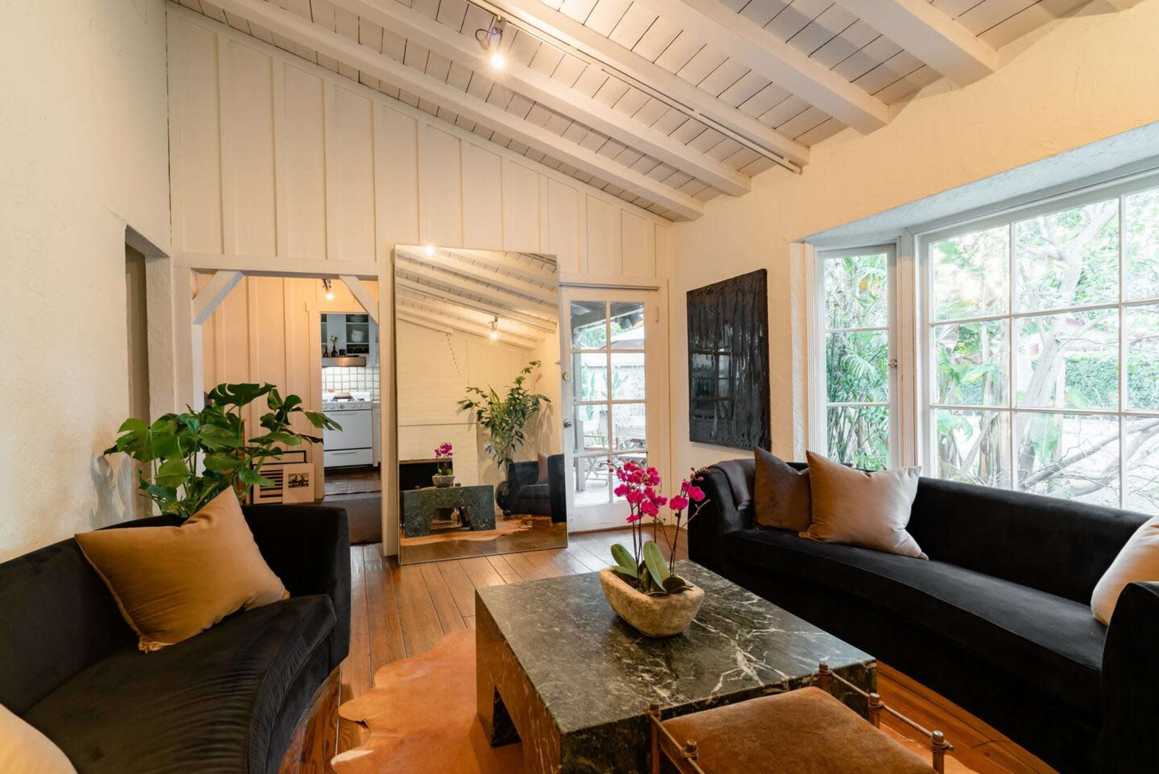 The image shows a living room with black sofas, a large mirror, a coffee table made of marble, and large windows letting in natural light.