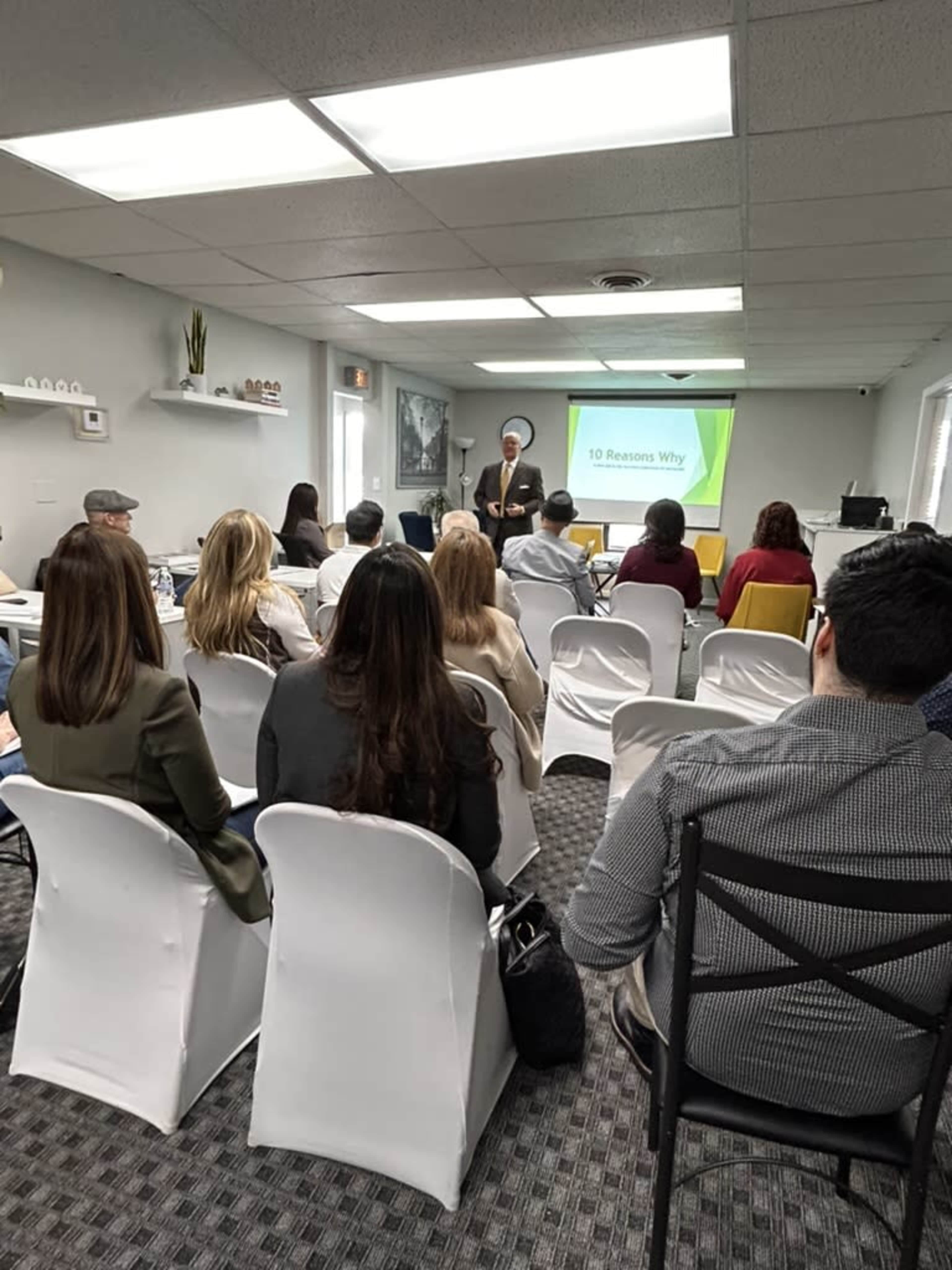 A speaker presents to an audience seated in a conference room, with a slideshow titled "10 Reasons Why" displayed on a screen.