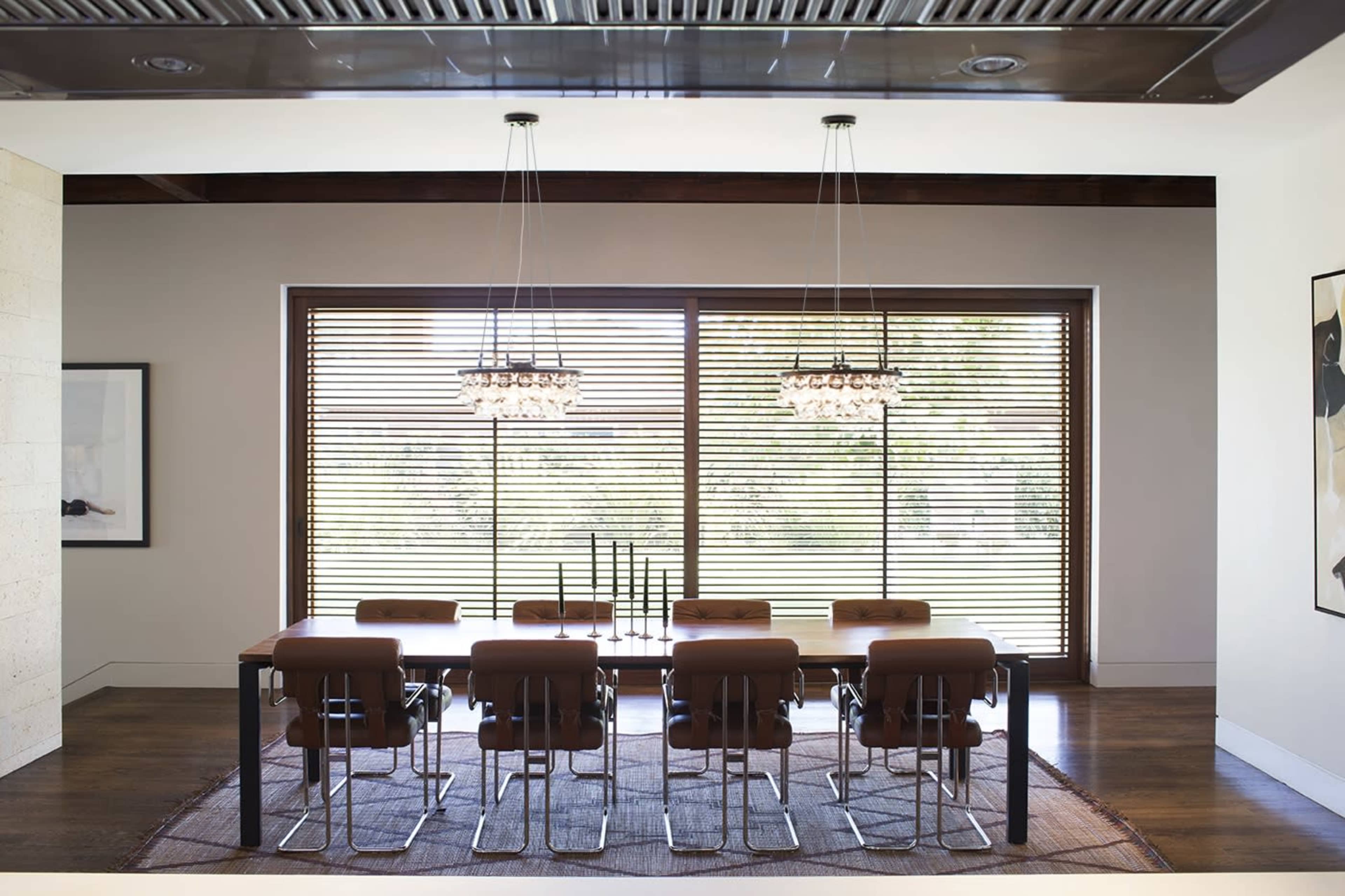 A modern dining room with a long wooden table surrounded by eight brown chairs, illuminated by two pendant lights and featuring large windows with horizontal blinds.