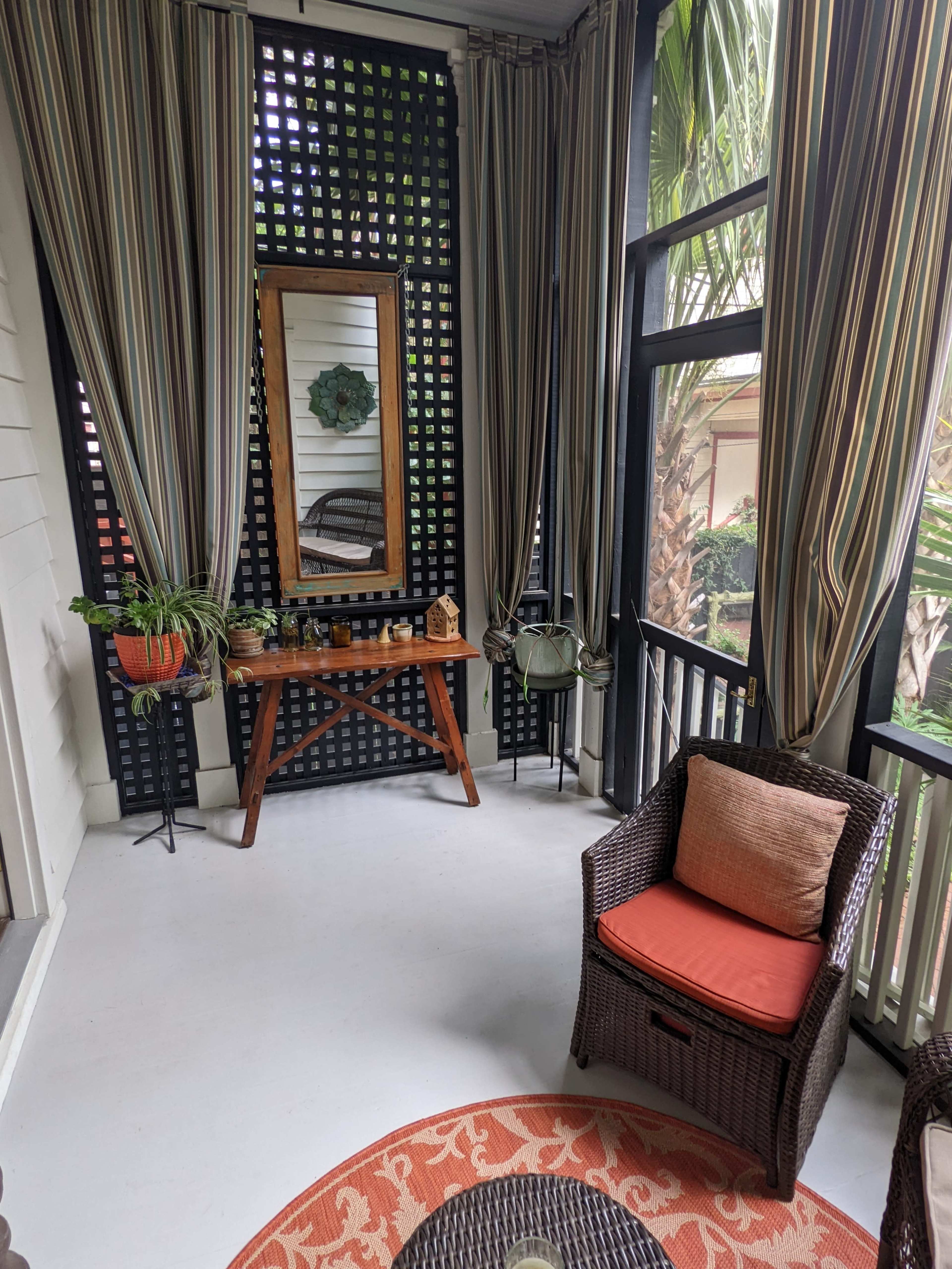A screened-in porch featuring wicker furniture, a wooden table, and decorative curtains, with greenery visible outside.