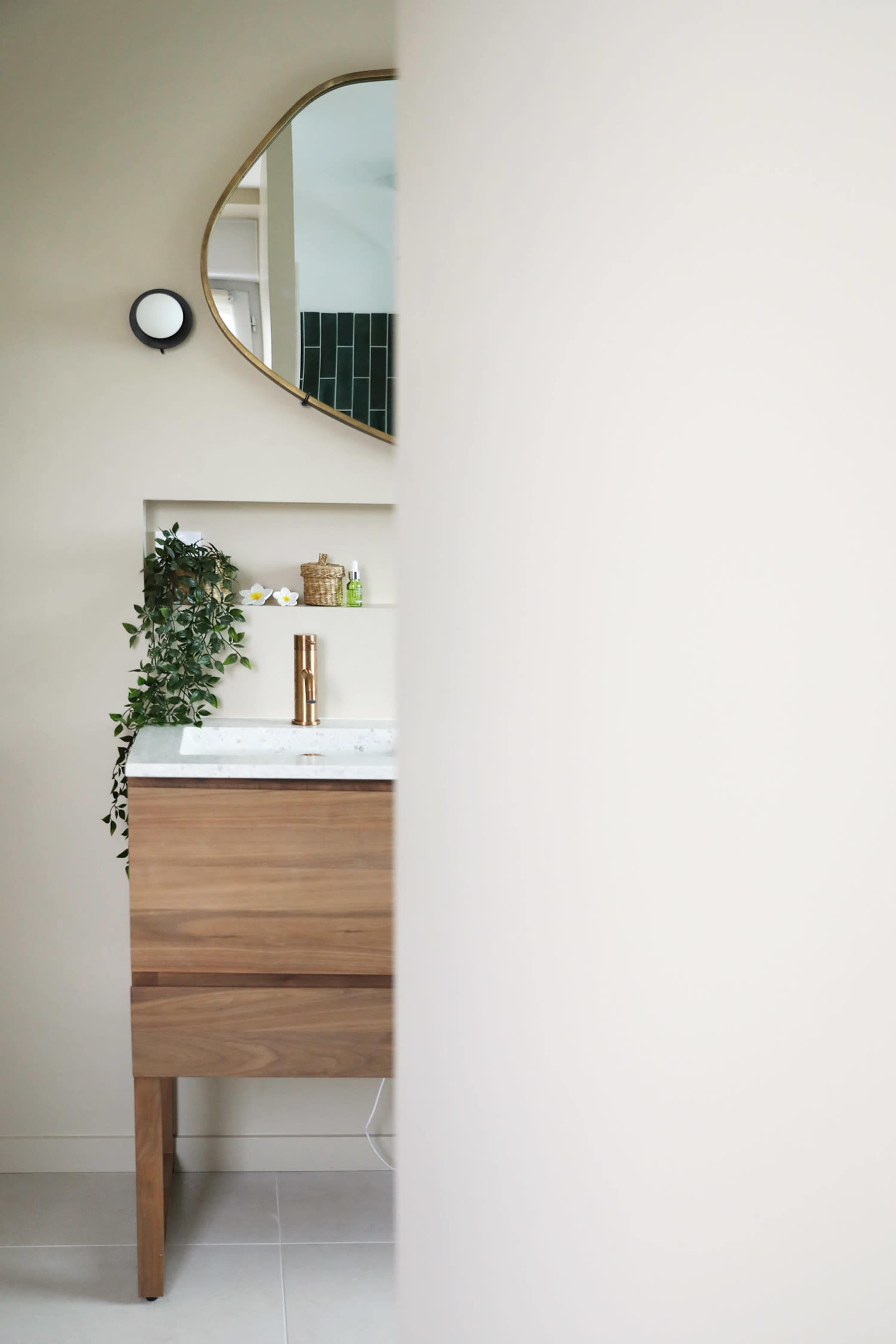 The image shows a bathroom with a wooden vanity, a round mirror above it, and green plants on the wall.