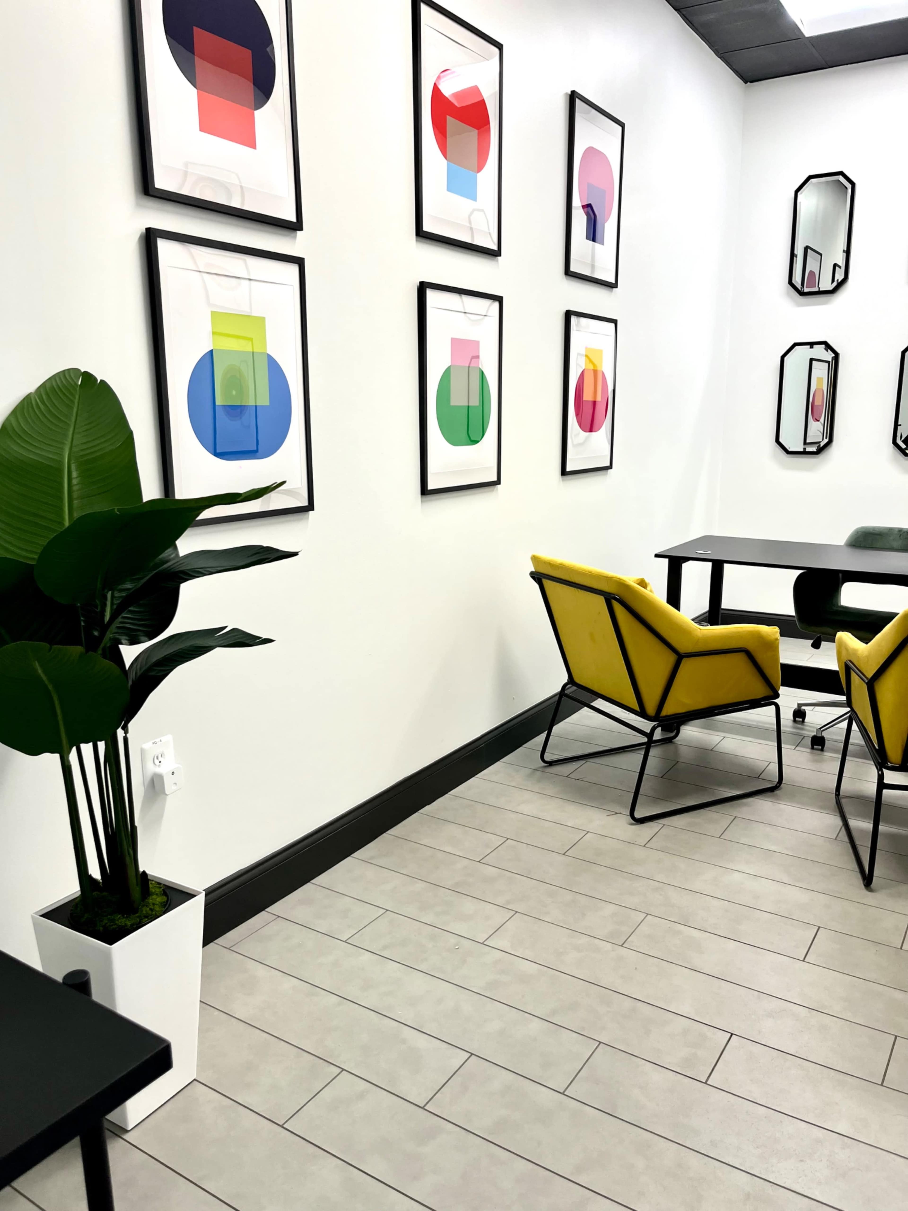 The image shows a modern office space with yellow chairs, a black table, a potted plant, and framed abstract art on the walls.