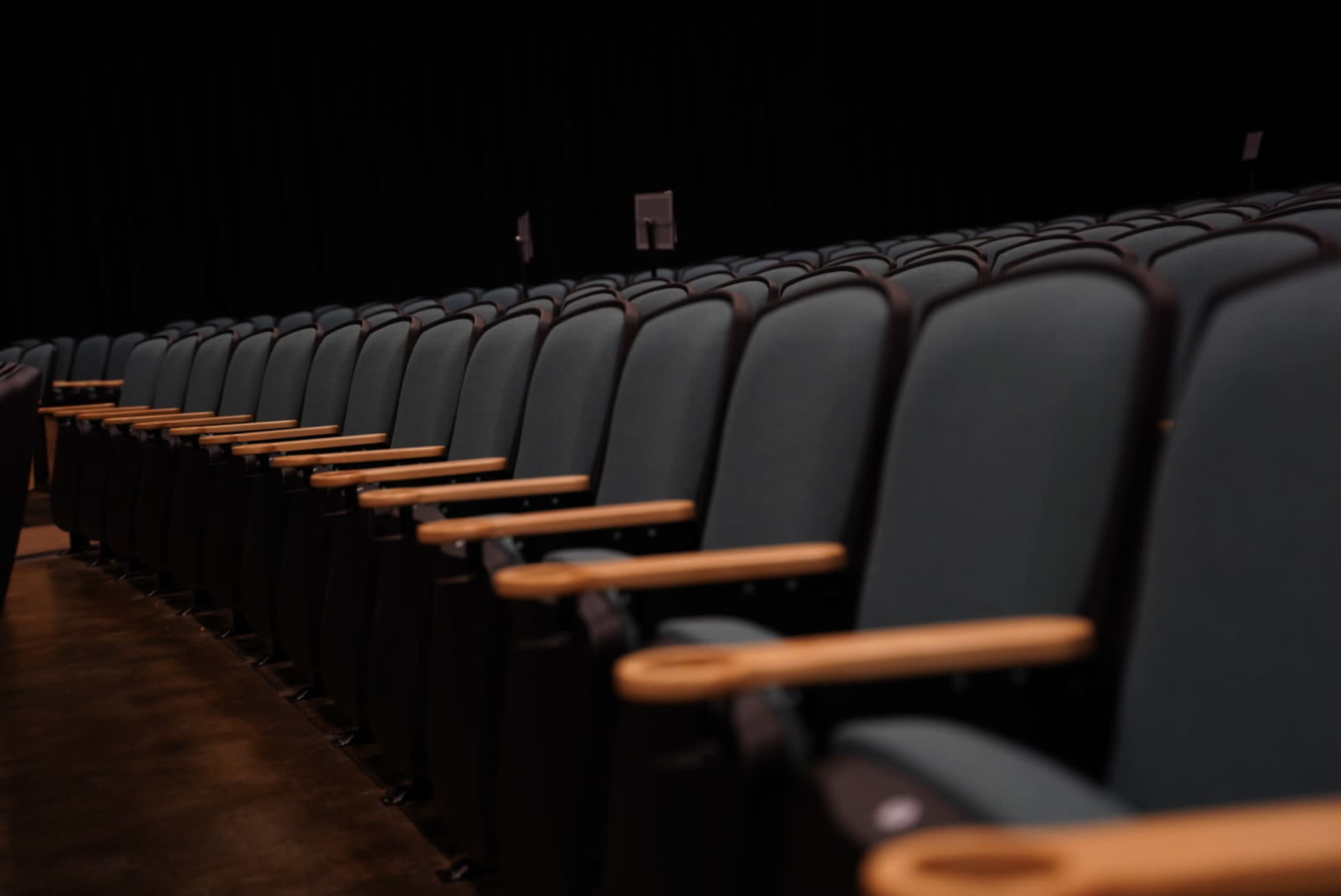 A row of empty theater seats with wooden armrests is lined up in a darkened auditorium.