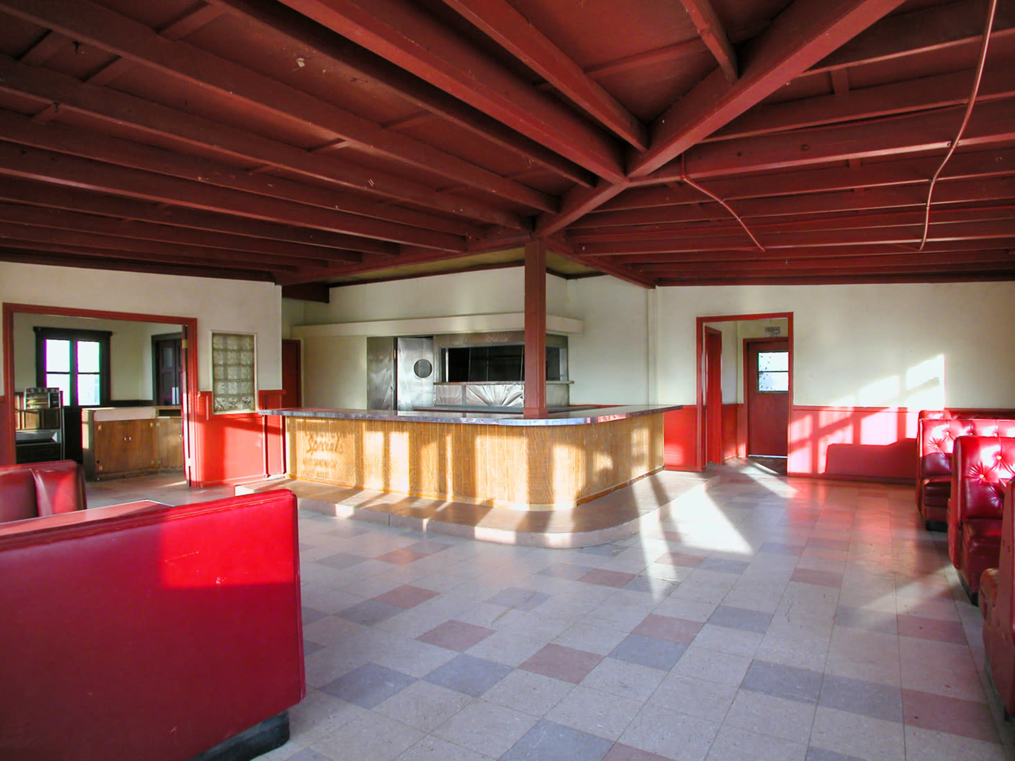 The image shows an empty room with red furniture and wooden beams, featuring a bar counter and a kitchen area in the background.