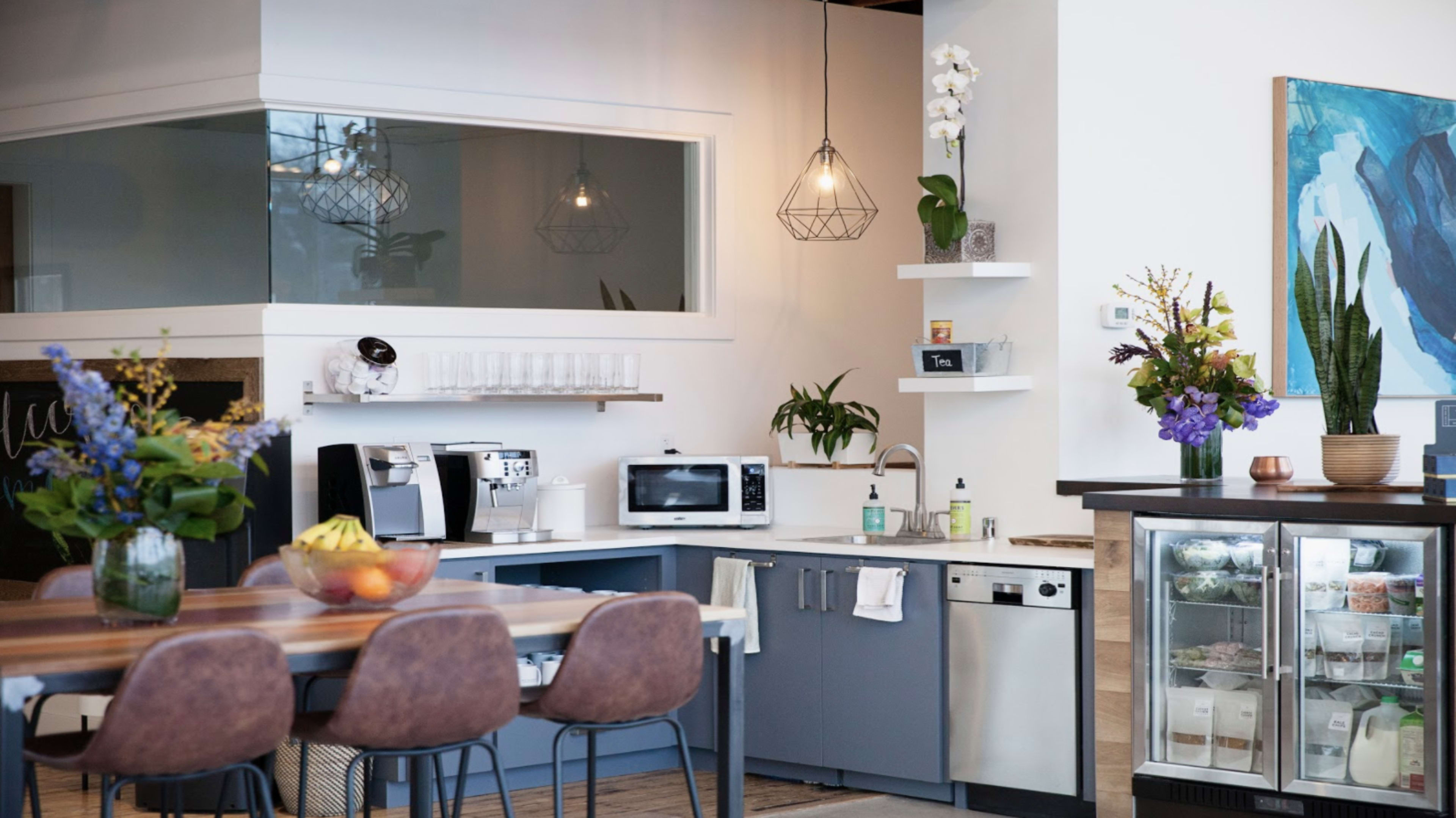 A modern kitchen area features a dining table with brown chairs, a coffee machine, a microwave, and a refrigerator, along with decorative plants and flowers.