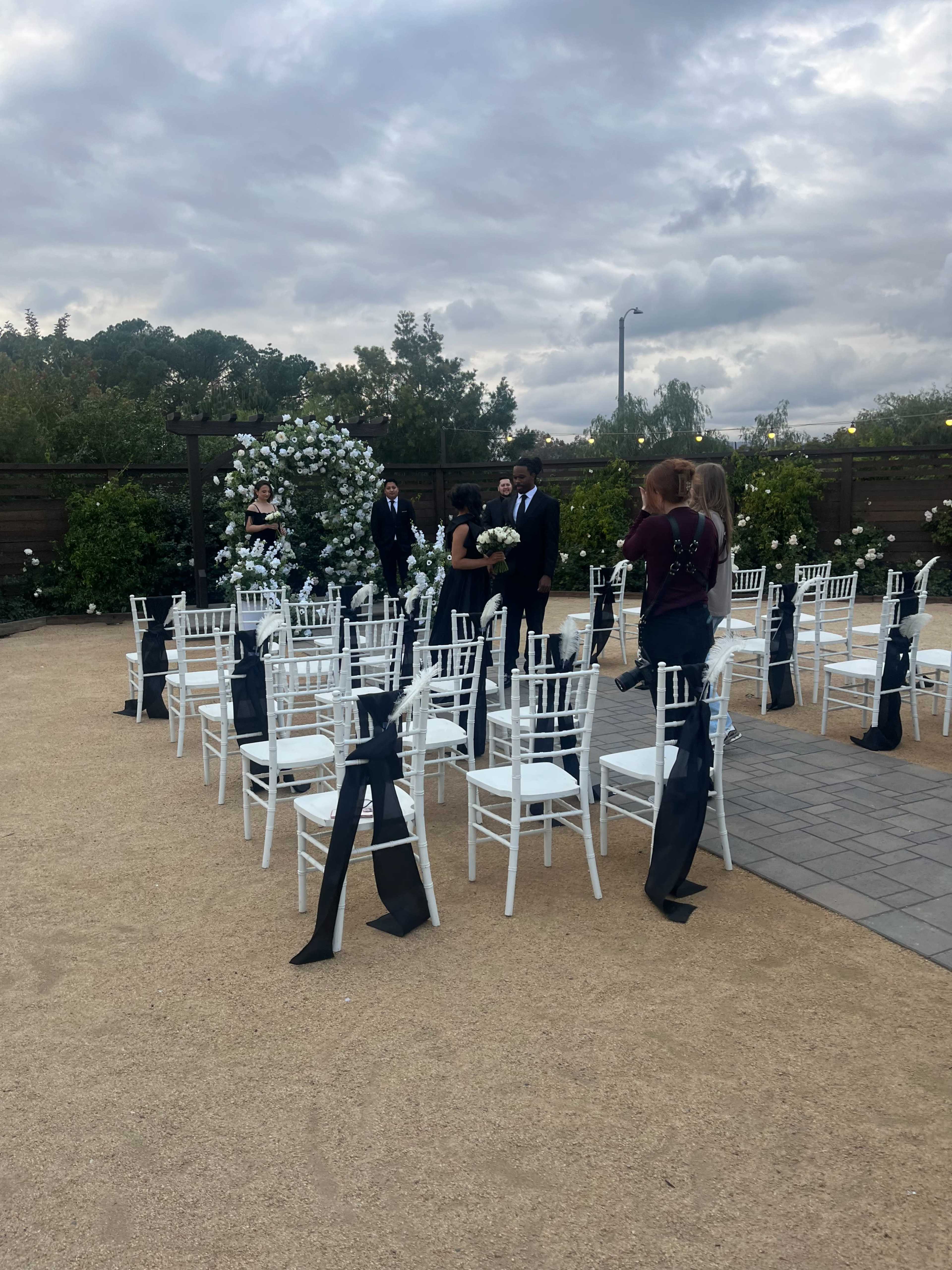 A wedding ceremony setup features rows of white chairs adorned with black sashes, surrounded by greenery under an overcast sky.