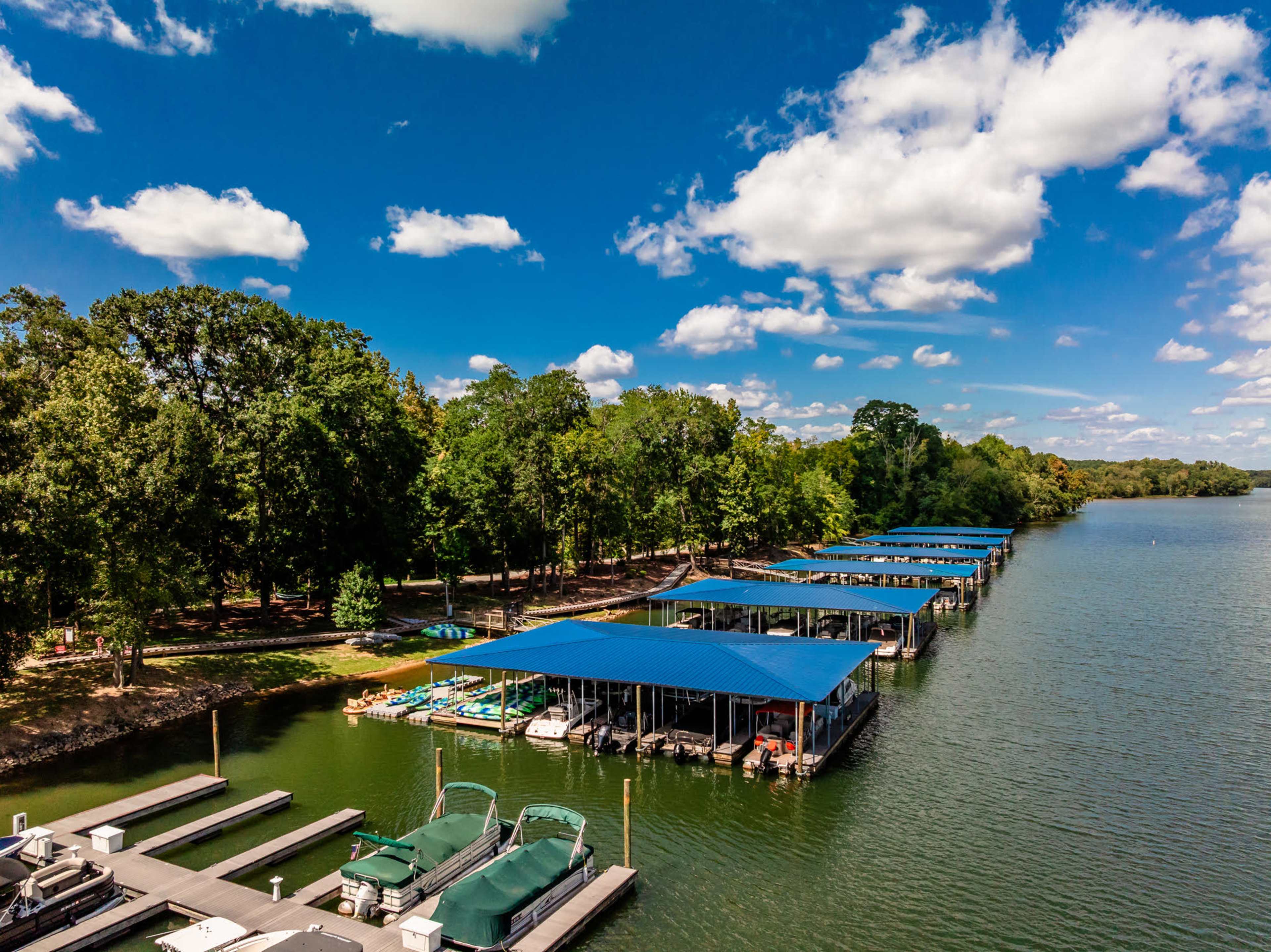 The image shows a marina on a calm lake with several covered boat docks and lush green trees lining the shore under a partly cloudy sky.