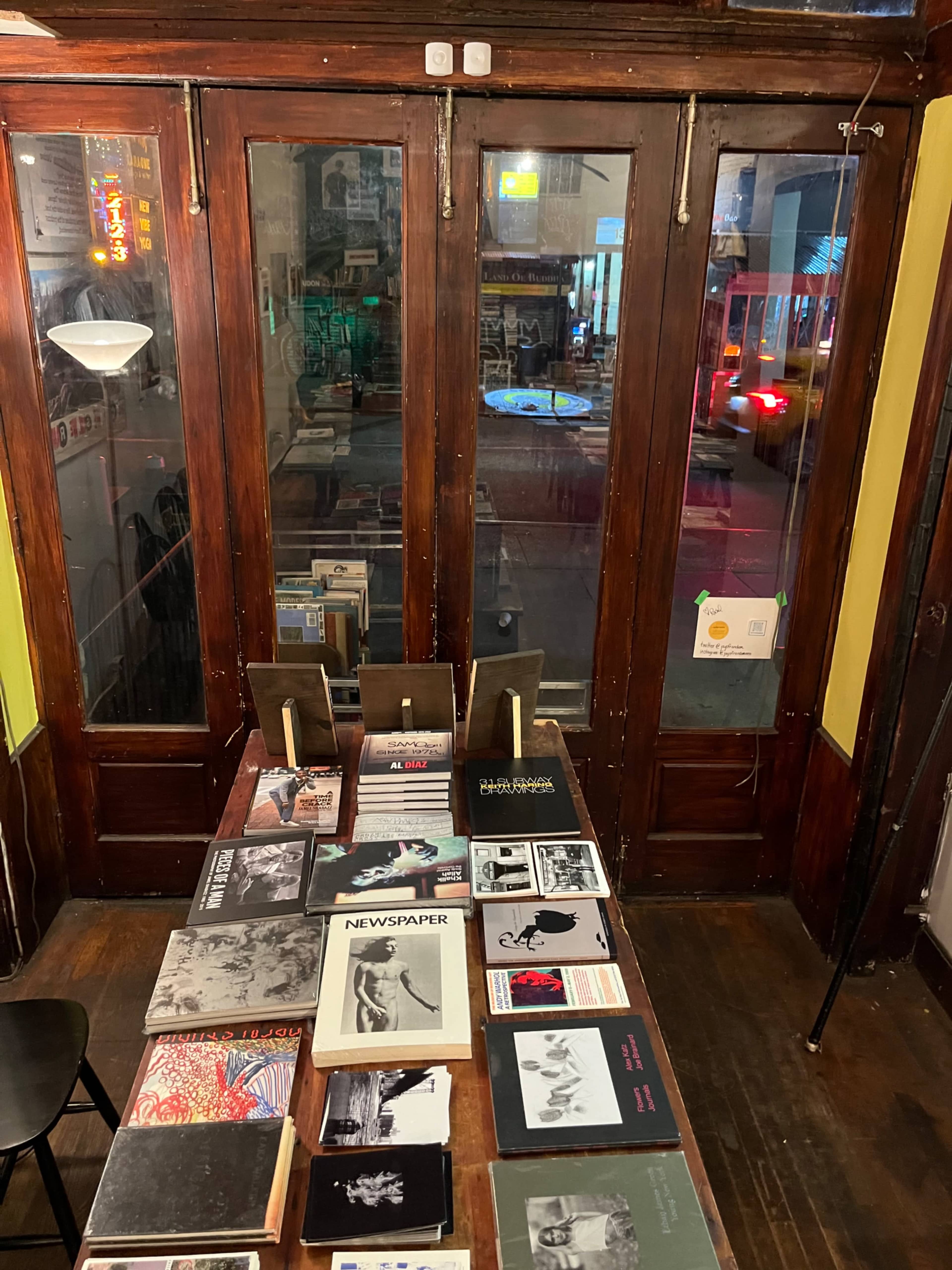 A wooden table is set with various books and magazines in front of large glass doors, revealing a street scene outside.