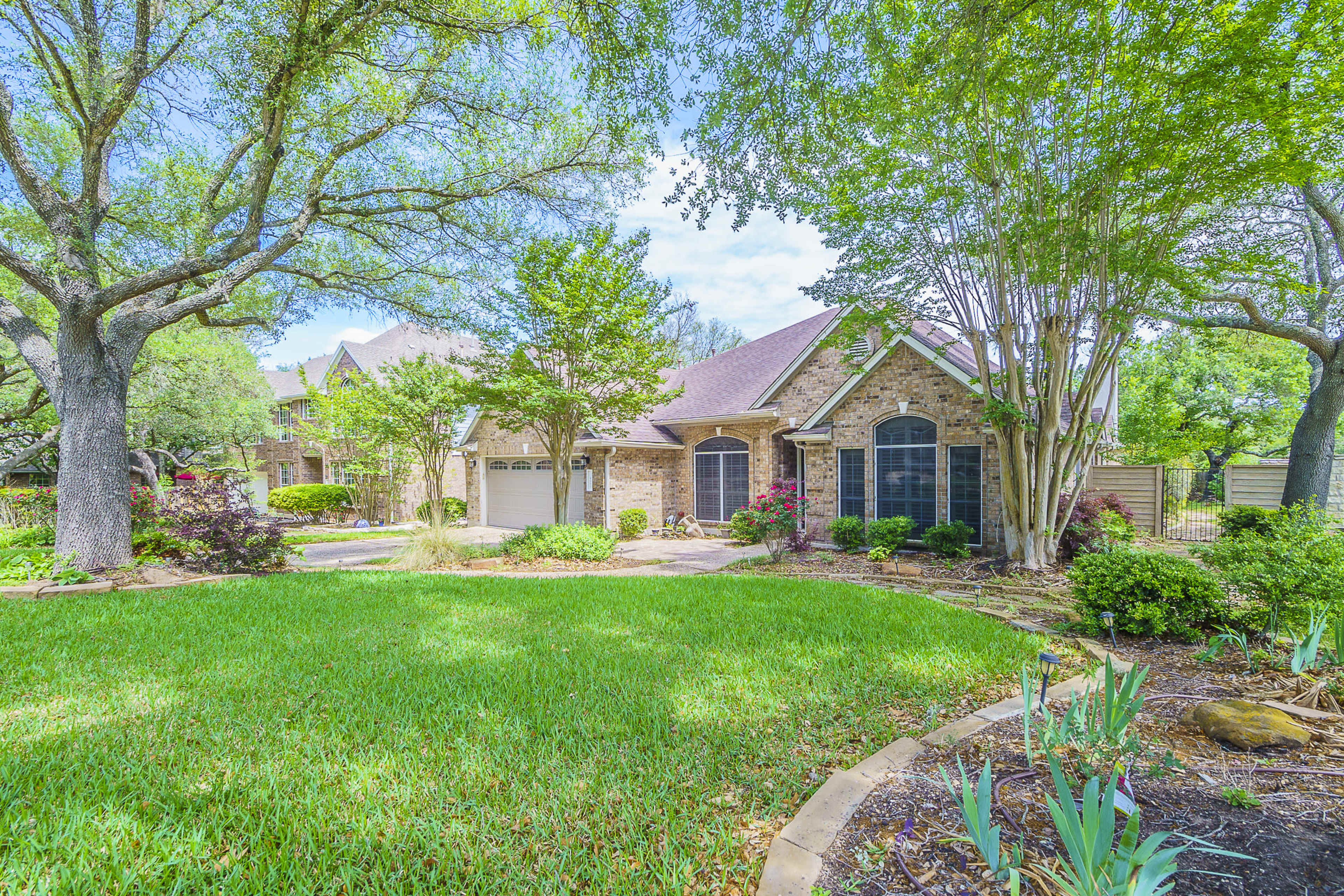 A well-maintained brick house with a landscaped front yard featuring green grass, trees, and flowering plants.