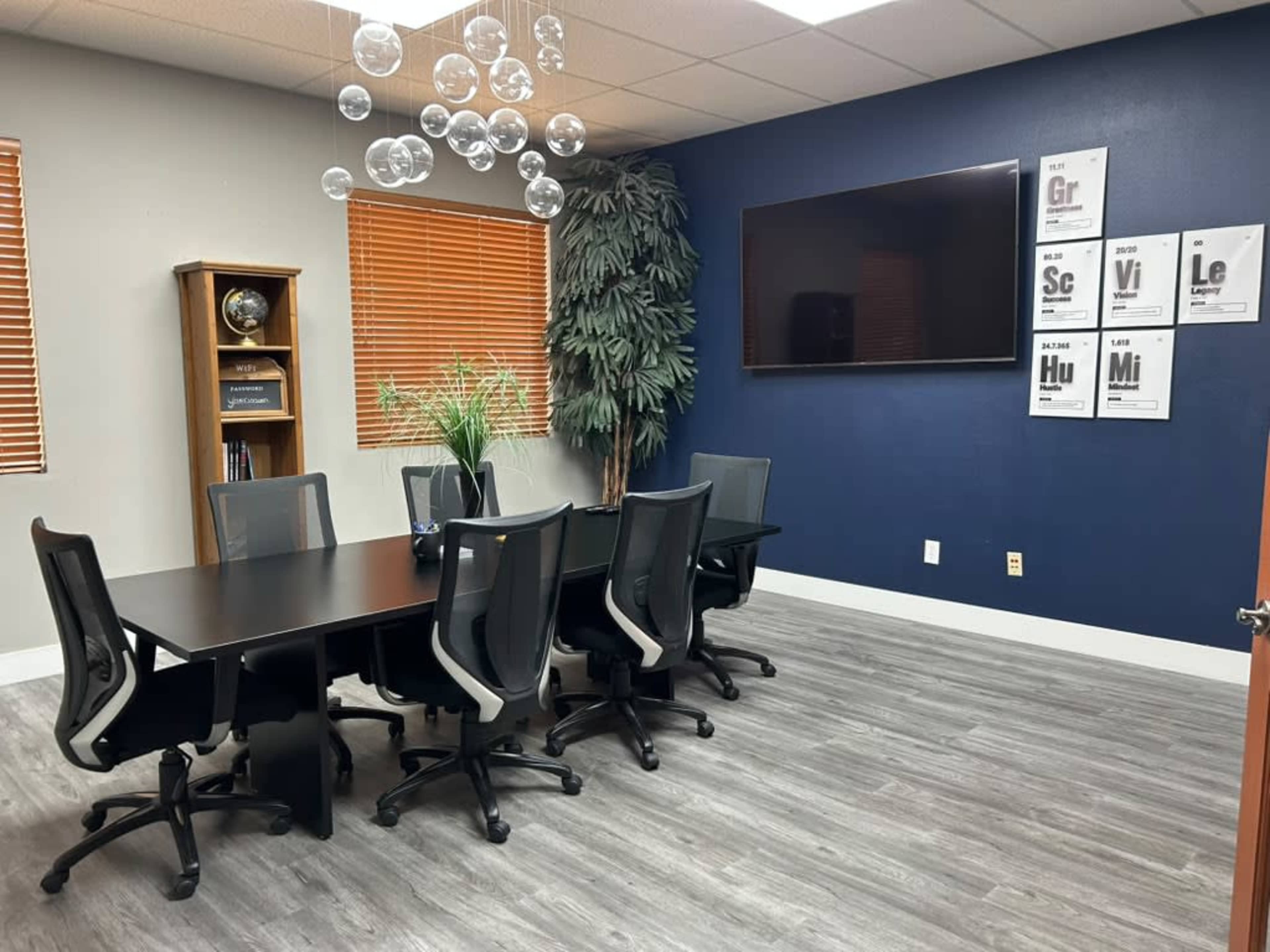 The image shows a modern conference room with a large table surrounded by eight chairs, a wall-mounted TV, and decorative elements including a potted plant and bubble-shaped light fixtures.