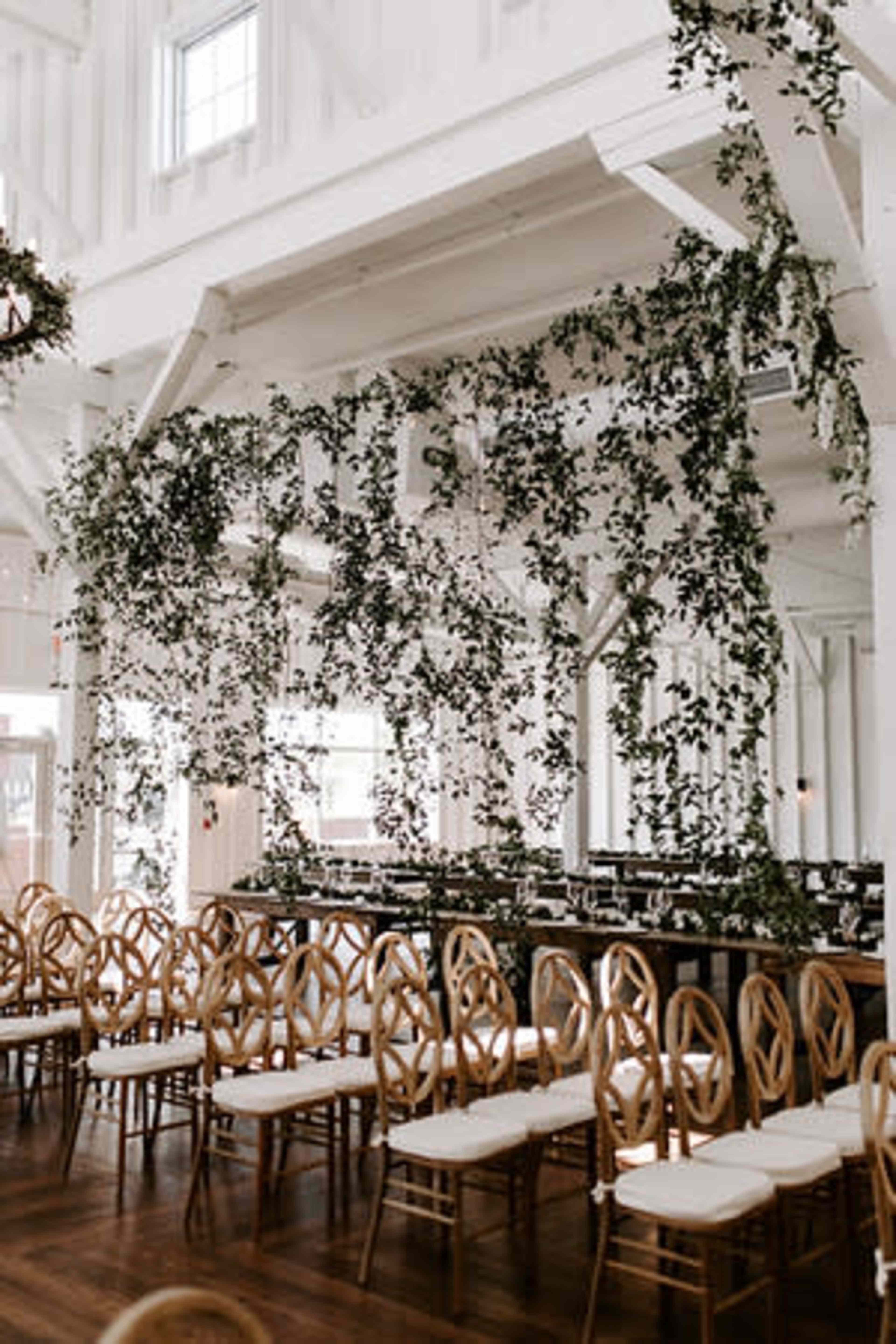 A spacious indoor venue with wooden chairs arranged in rows, adorned with hanging greenery above a dining area.
