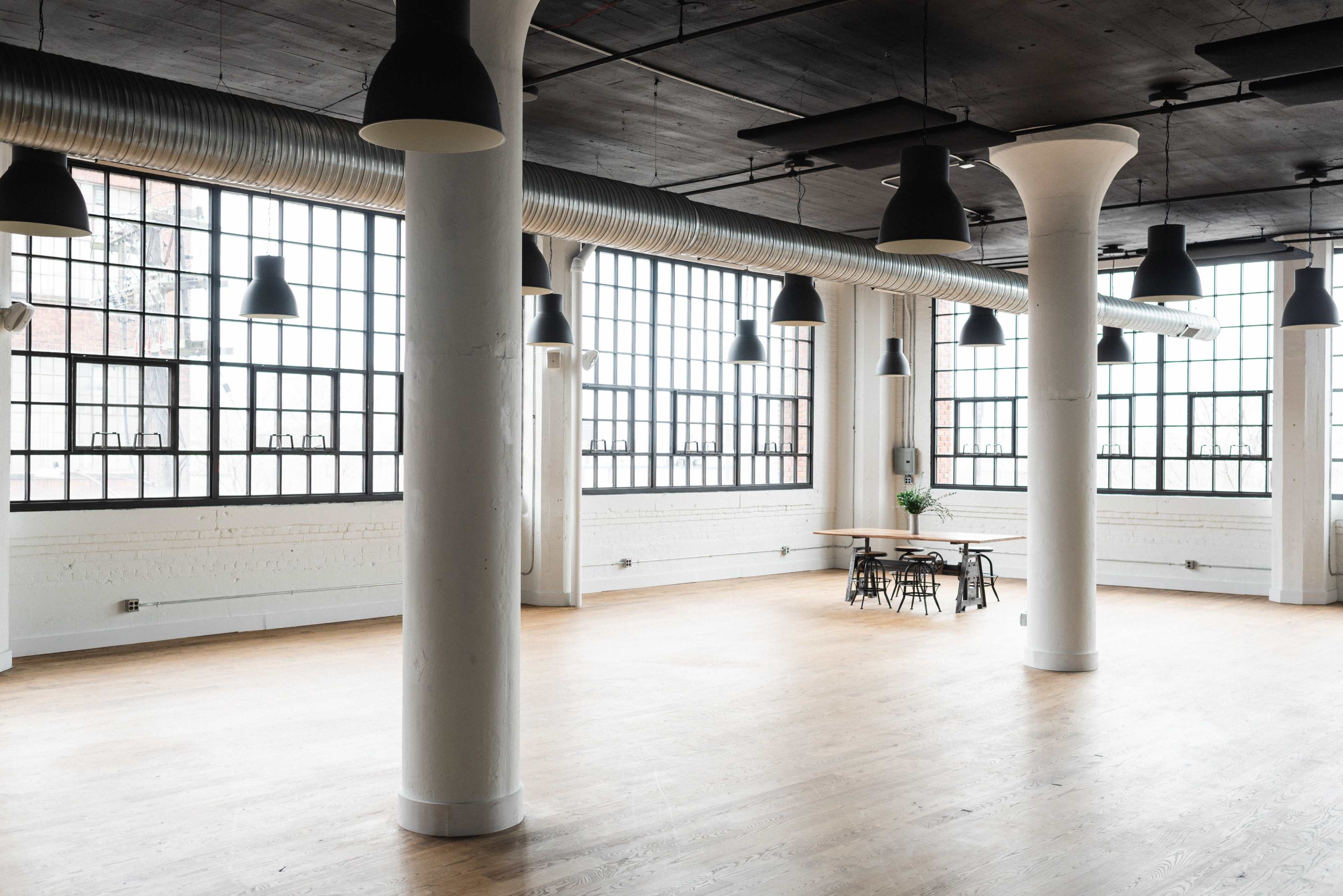 A spacious, industrial-style room with large windows, high ceilings, and a wooden floor, featuring several columns and a dining table with chairs.