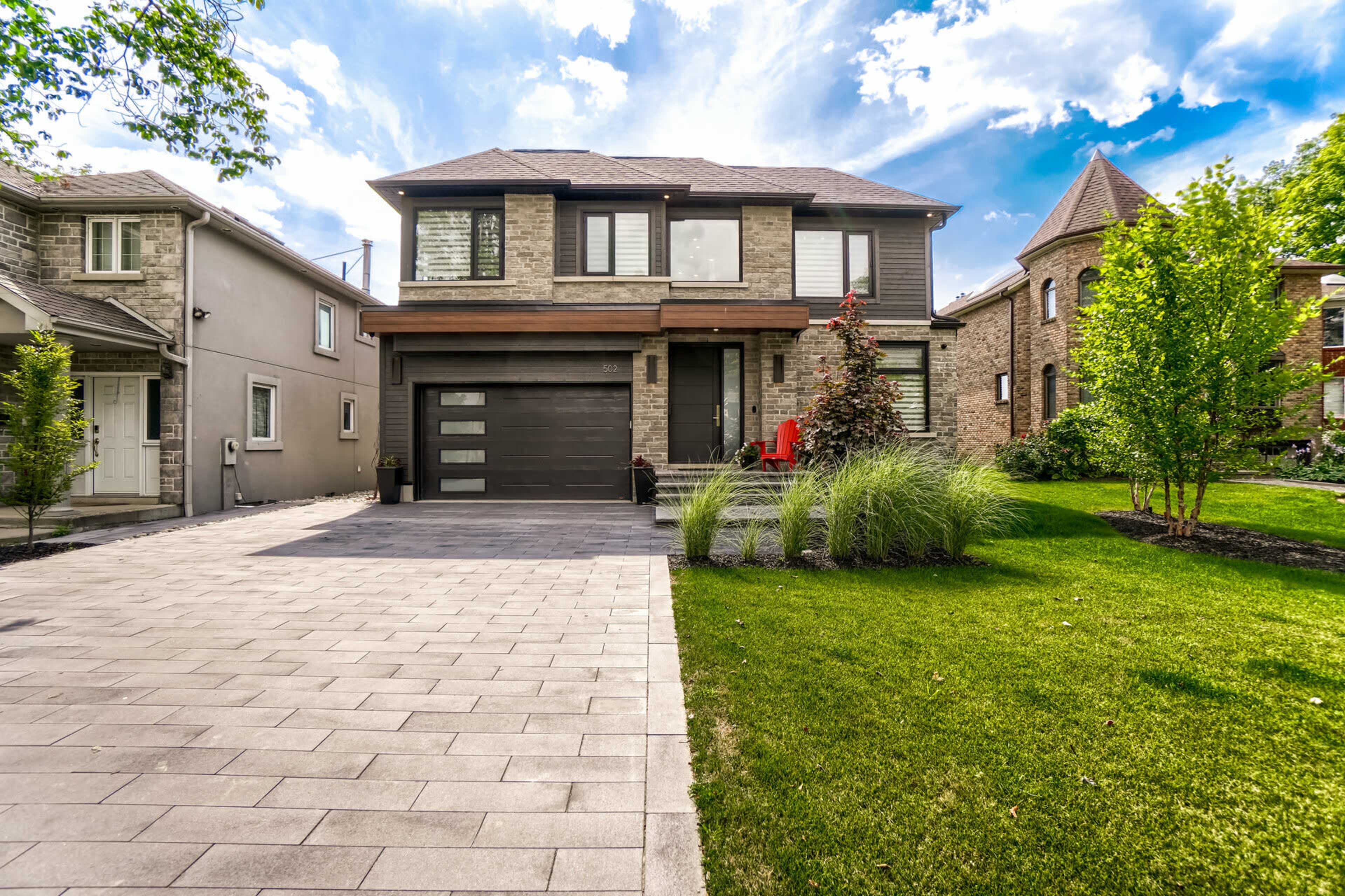 A two-story brick house with a grey garage door is surrounded by a landscaped yard featuring grass, shrubs, and trees.