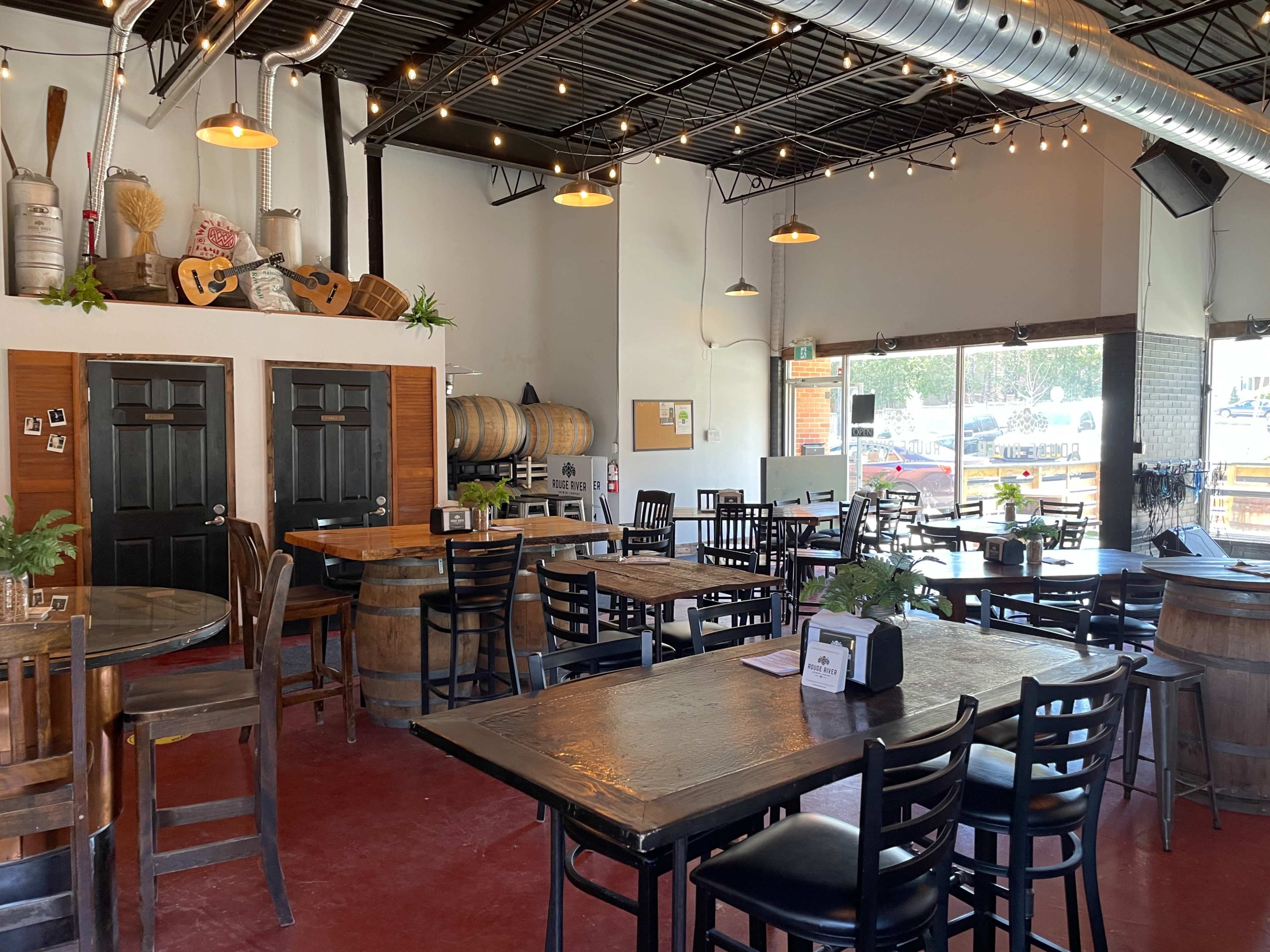 An interior of a casual restaurant featuring wooden tables, black chairs, decorative plants, and barrels lined against the walls.
