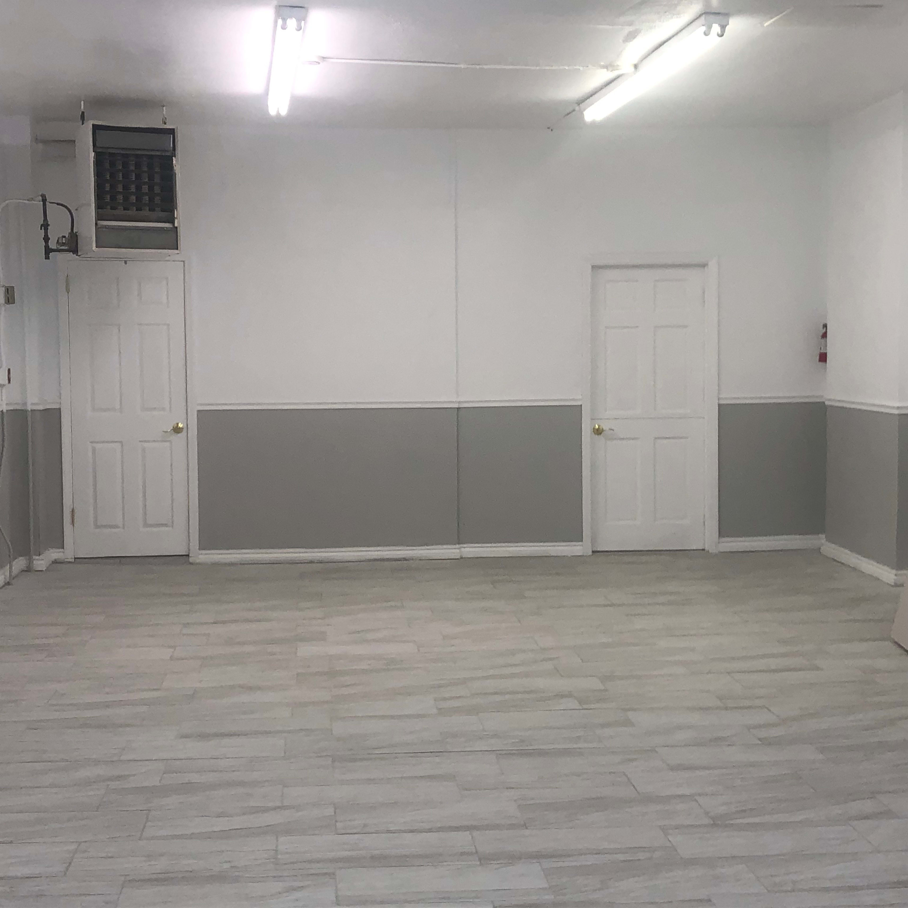 The image shows a spacious, empty room with gray and white walls, two doors, and fluorescent lighting.