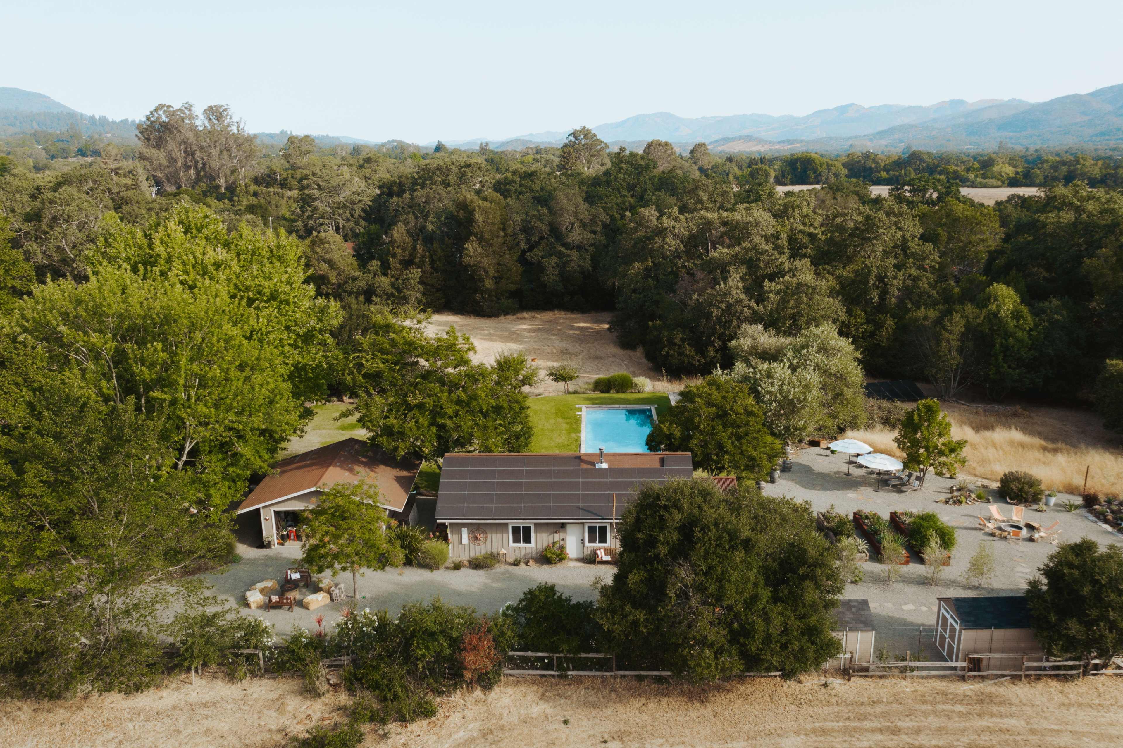 A rural property featuring a house with a solar panel roof, a swimming pool, and various trees and open land surrounding it.