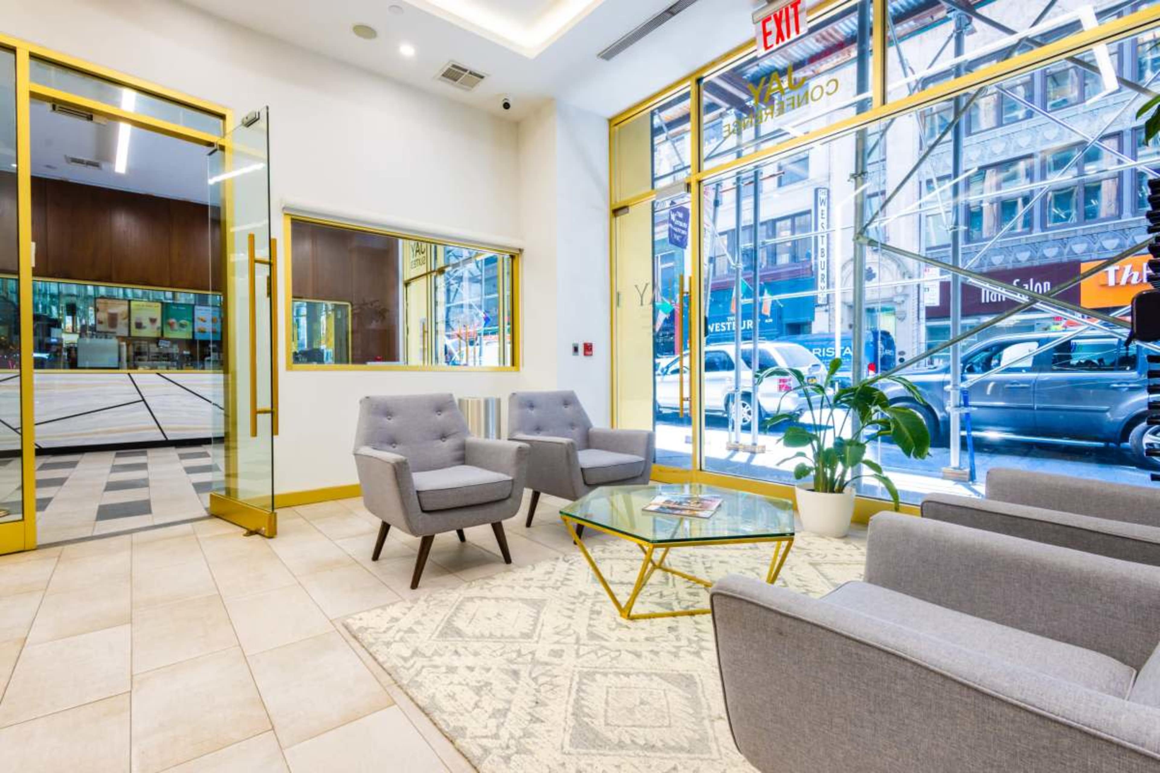 The image shows a modern lobby with gray armchairs, a glass coffee table, and large windows facing a bustling street.