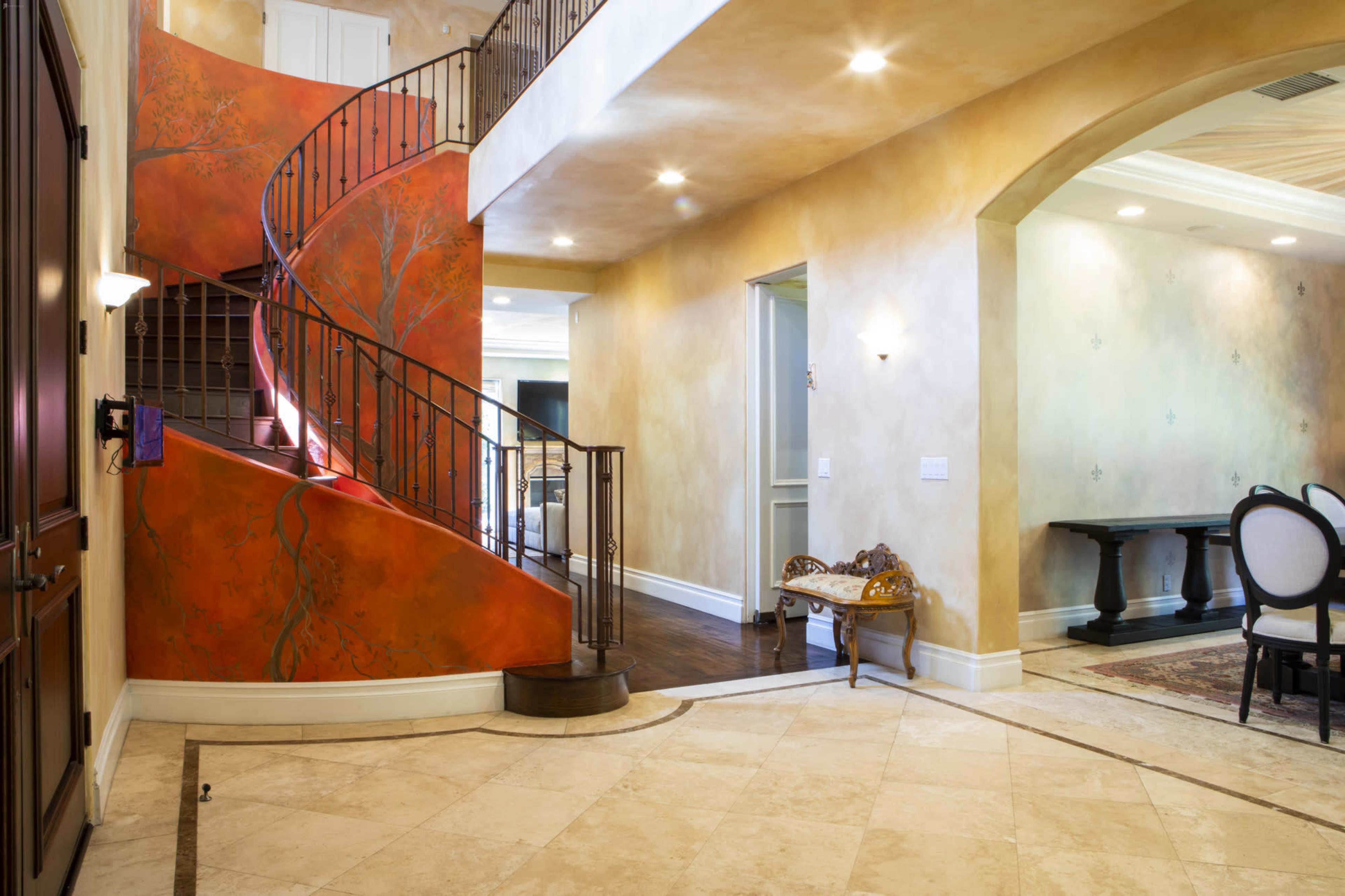 A two-story foyer featuring a curved staircase with a red and tree-decorated wall and marble flooring.
