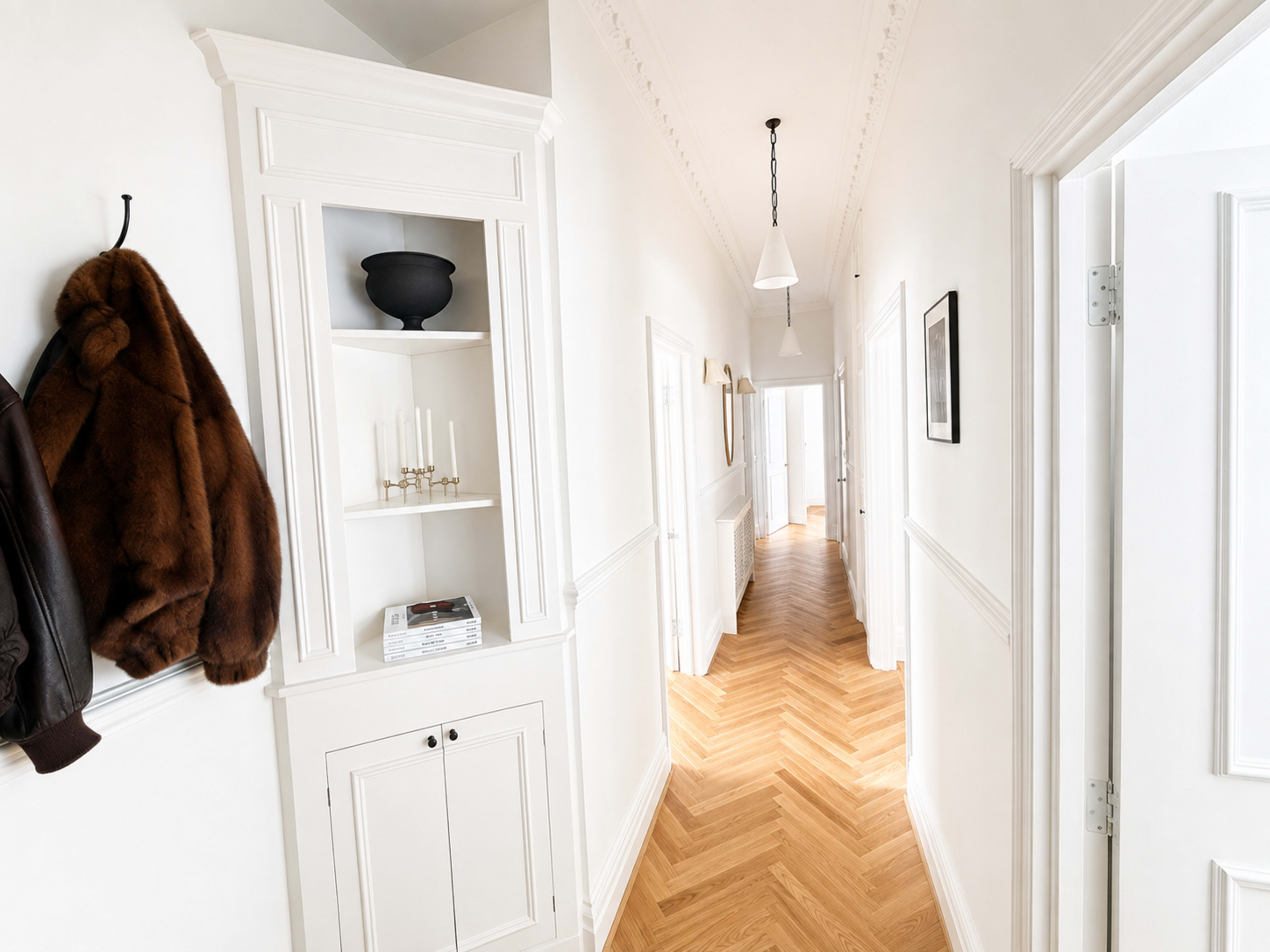 The image shows a bright, minimalist hallway with white walls, wooden flooring, and a shelf displaying a few items.