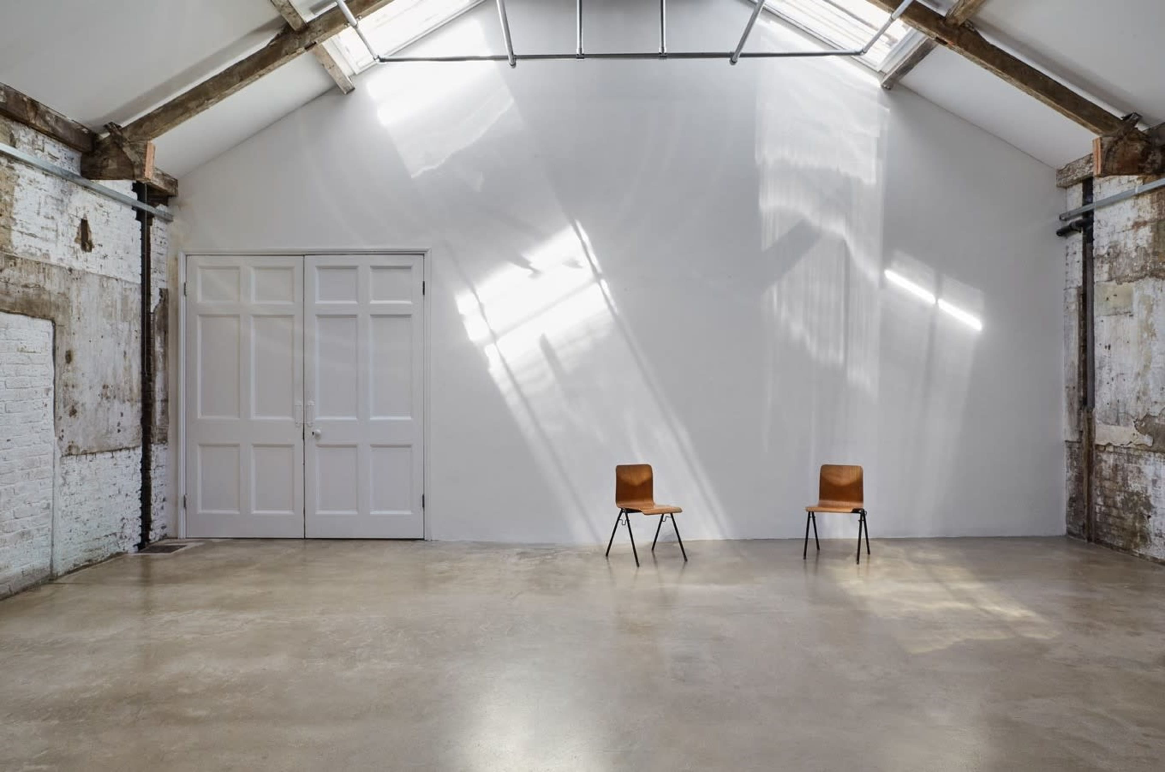 An empty industrial space features two chairs positioned on a polished concrete floor, with natural light streaming through a large window.