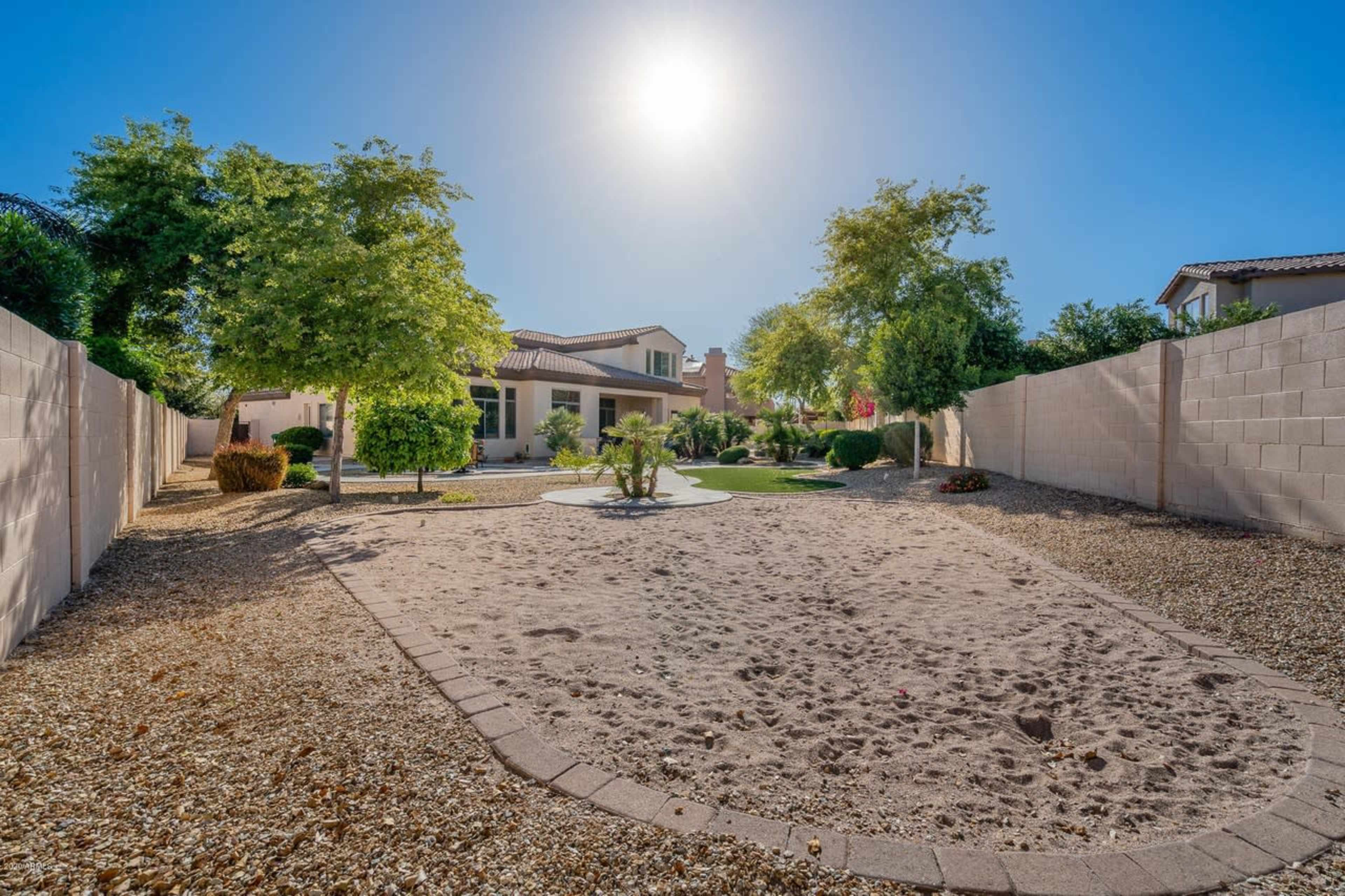 The image shows a spacious backyard with a sandy area surrounded by desert landscaping and a two-story house in the background under a bright sun.