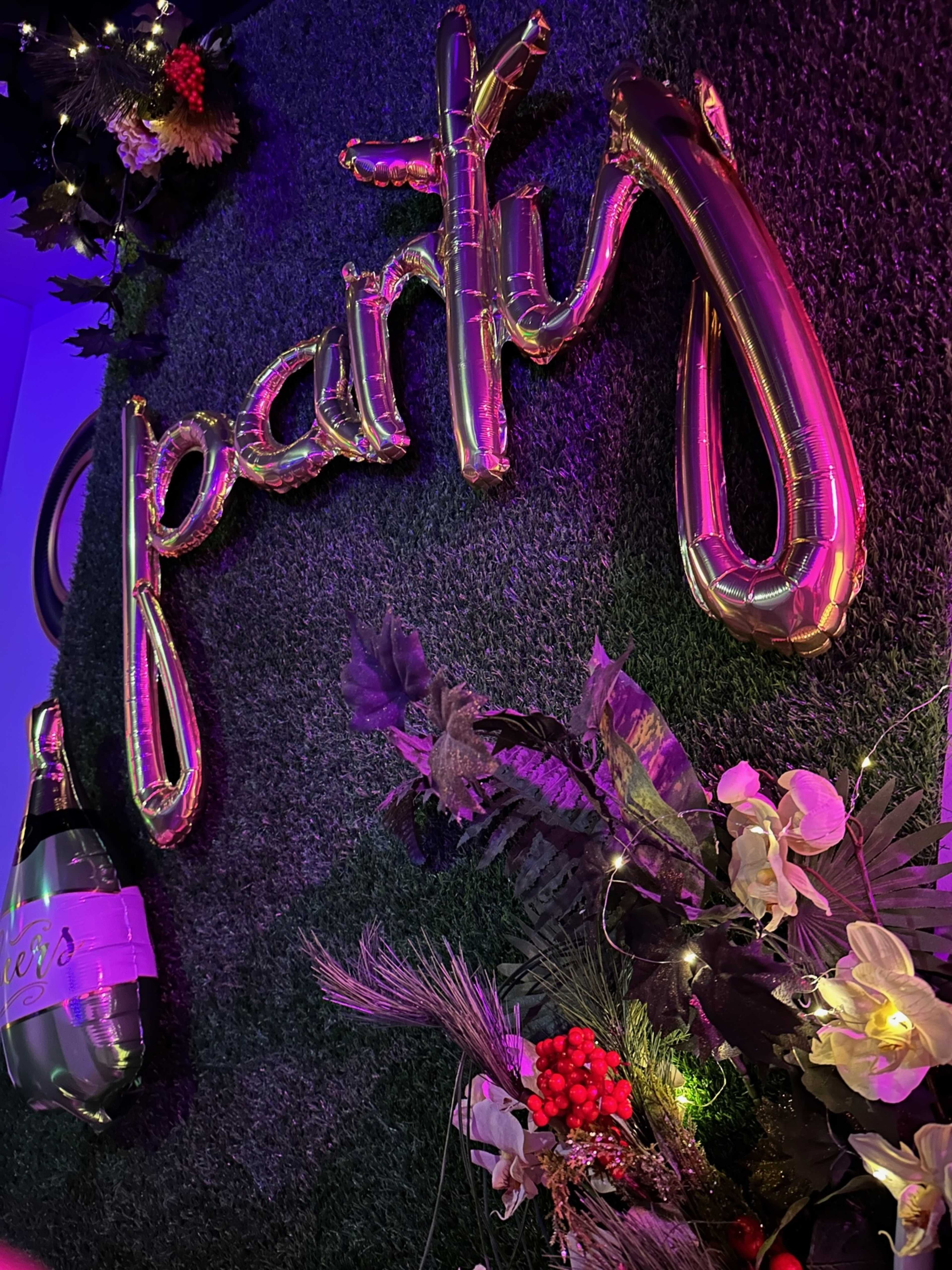 A decorative display featuring the word "party" formed with metallic balloons, surrounded by artificial flowers and a bottle of champagne on a grass-like backdrop.
