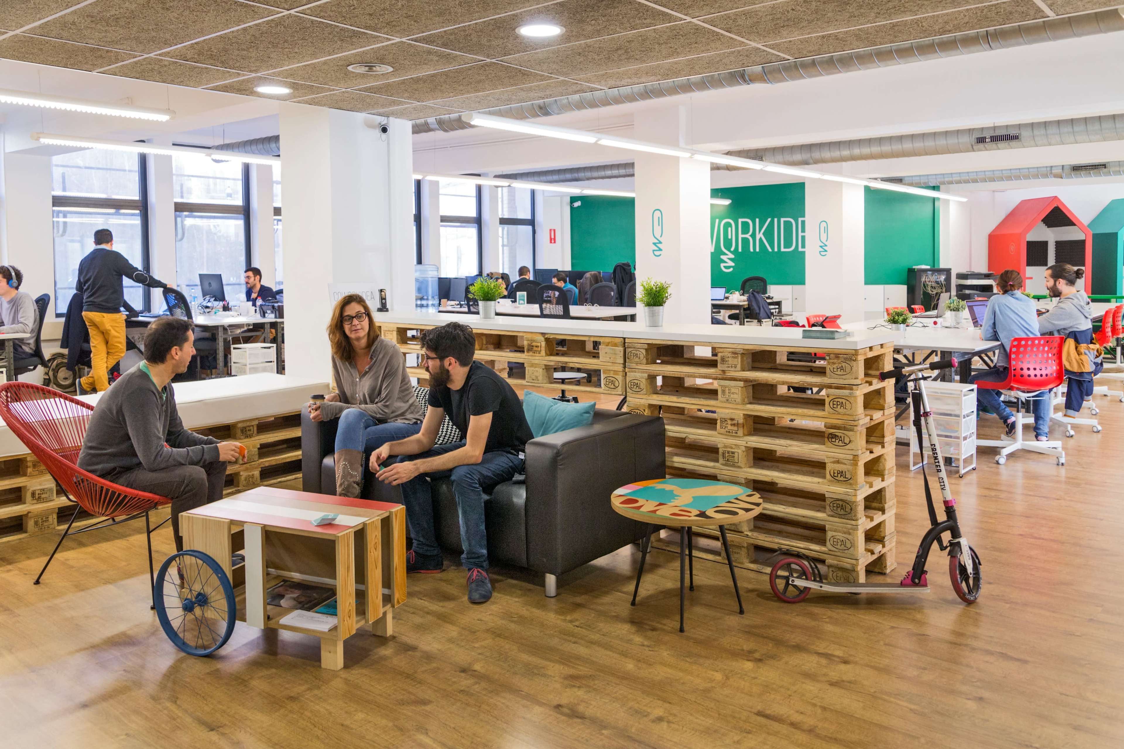 A modern office space features seating areas with pallet furniture, a scooter, and several workers engaged at desks in the background.