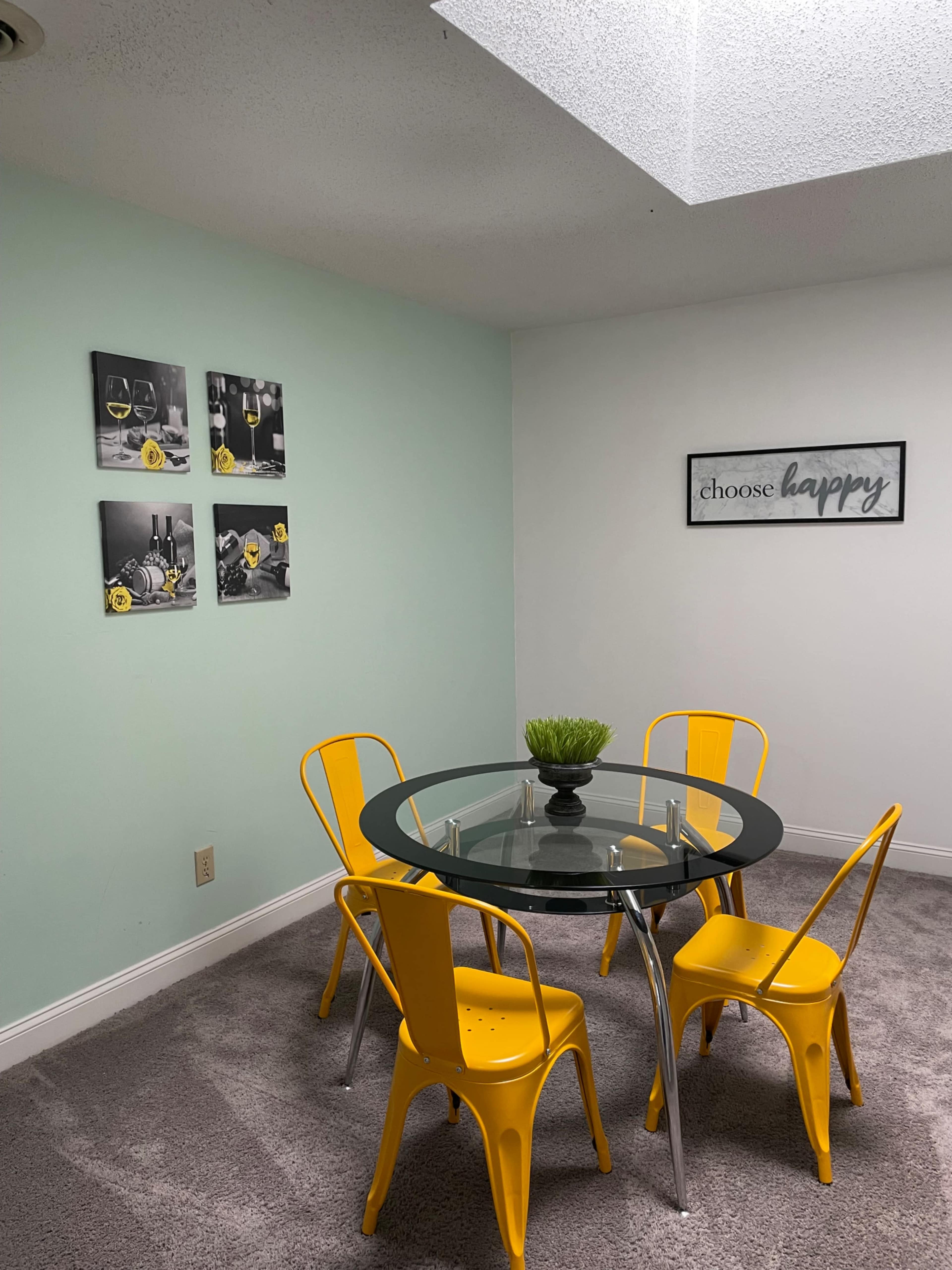 A round glass dining table is surrounded by four yellow chairs in a well-lit room with light green walls and decorative wall art.