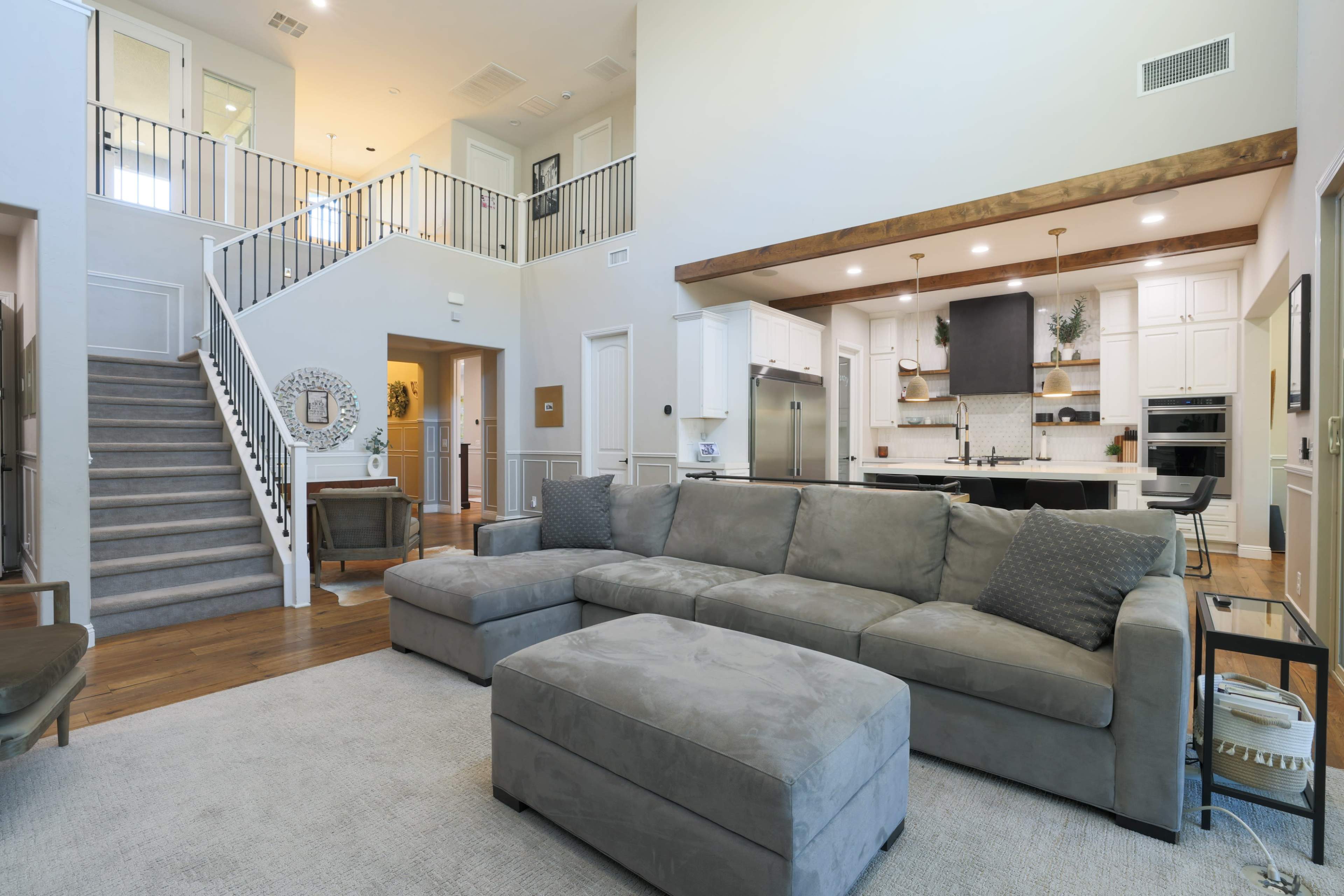 The image shows a modern living room with a large sectional sofa, an ottoman, and an open-concept kitchen and staircase in the background.