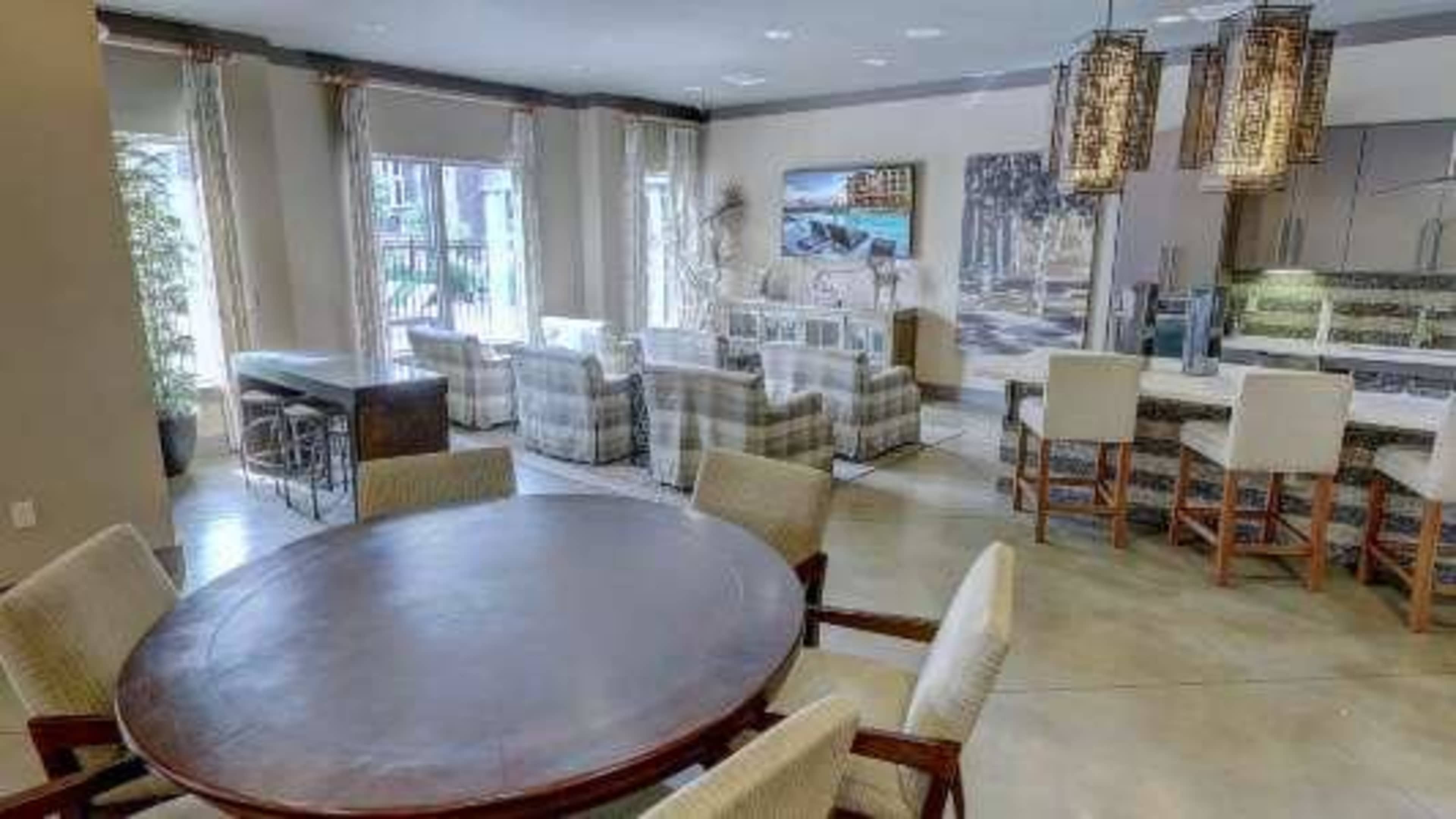 The image shows a modern open-concept living area with a dining table, upholstered chairs, and a kitchen with bar seating, surrounded by large windows and decorative light fixtures.