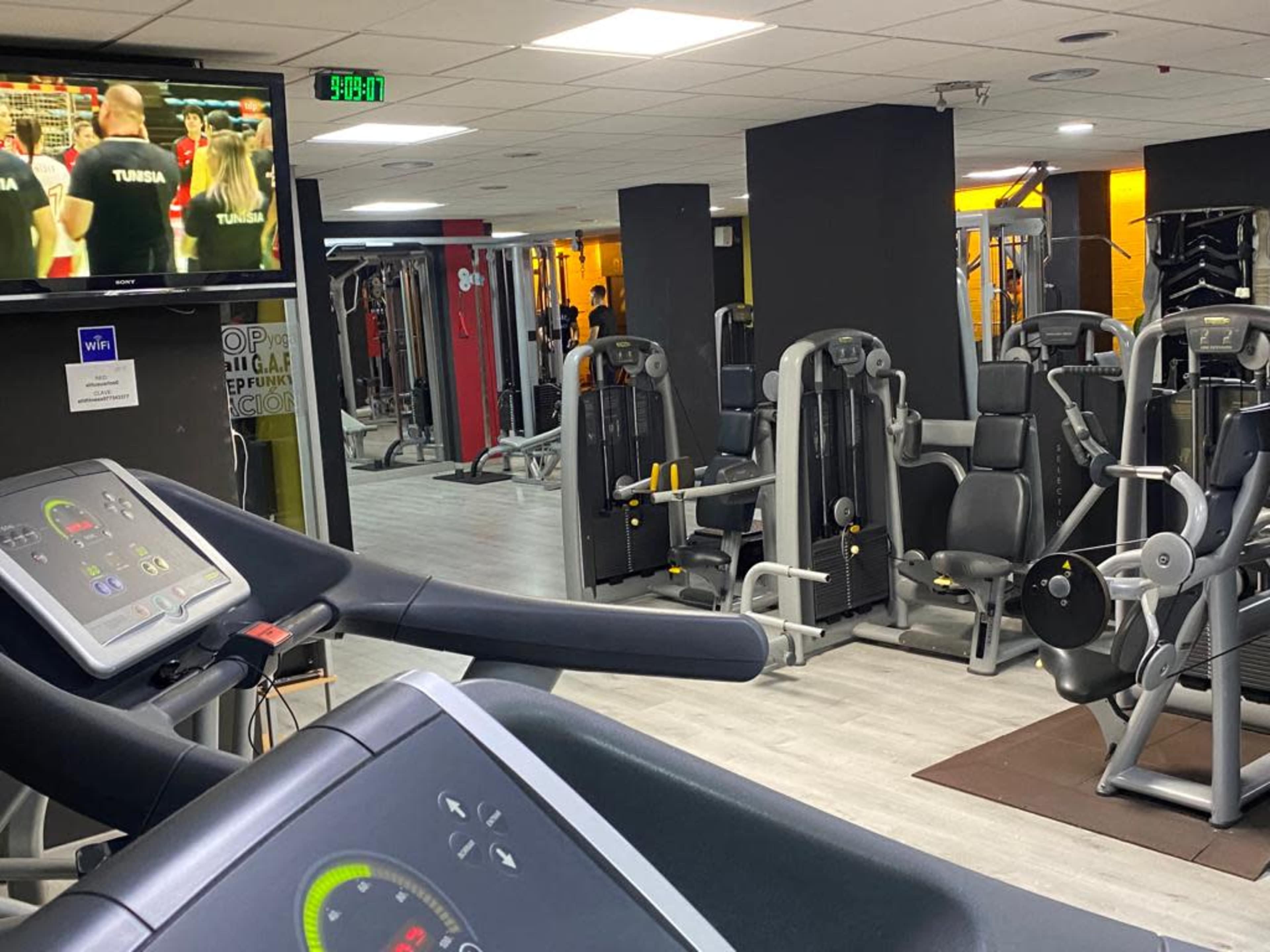 A modern gym interior with various fitness machines and equipment, including treadmills and weight training machines, along with a television displaying a sports event.