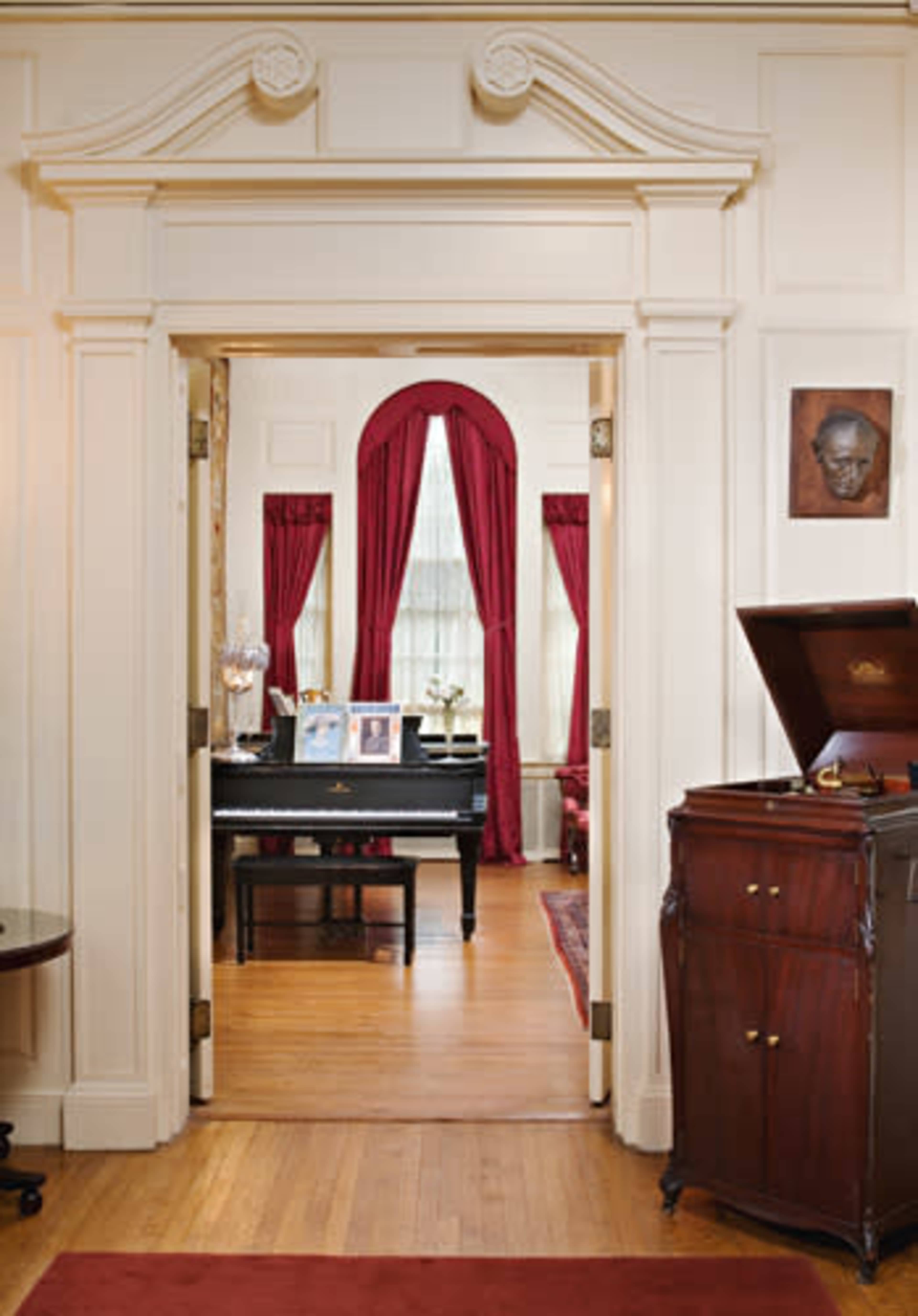 A doorway framed by decorative molding leads into a room with a piano, photographs on display, and rich red curtains.