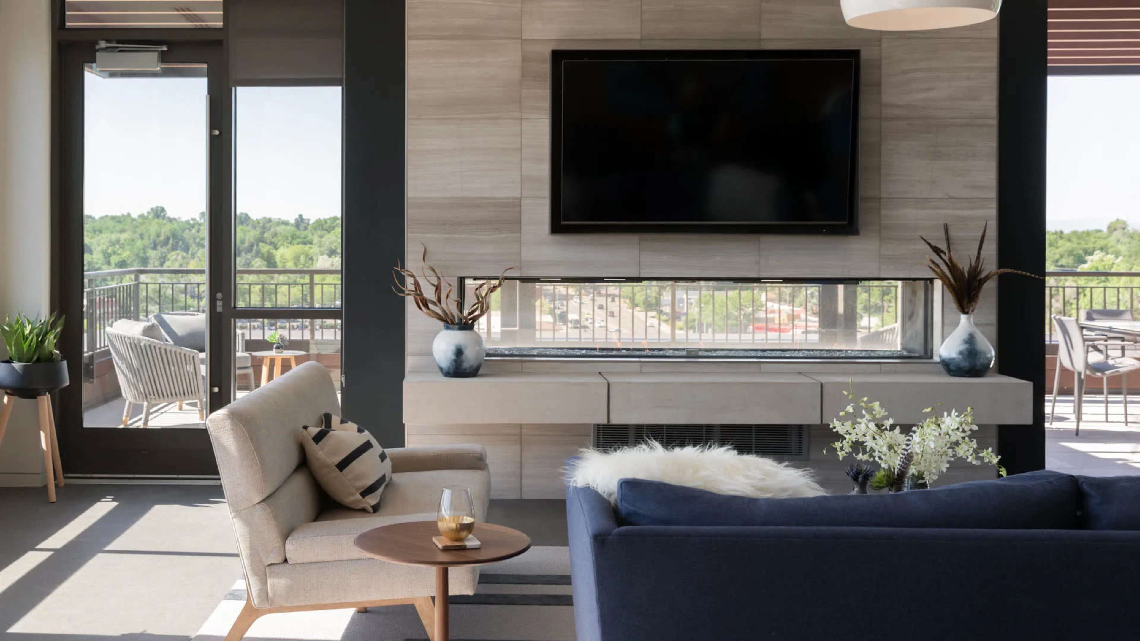 A modern living space features a wall-mounted television above a sleek console, with a comfortable sofa and a small table holding a drink, and large windows that overlook a greenery-filled view.