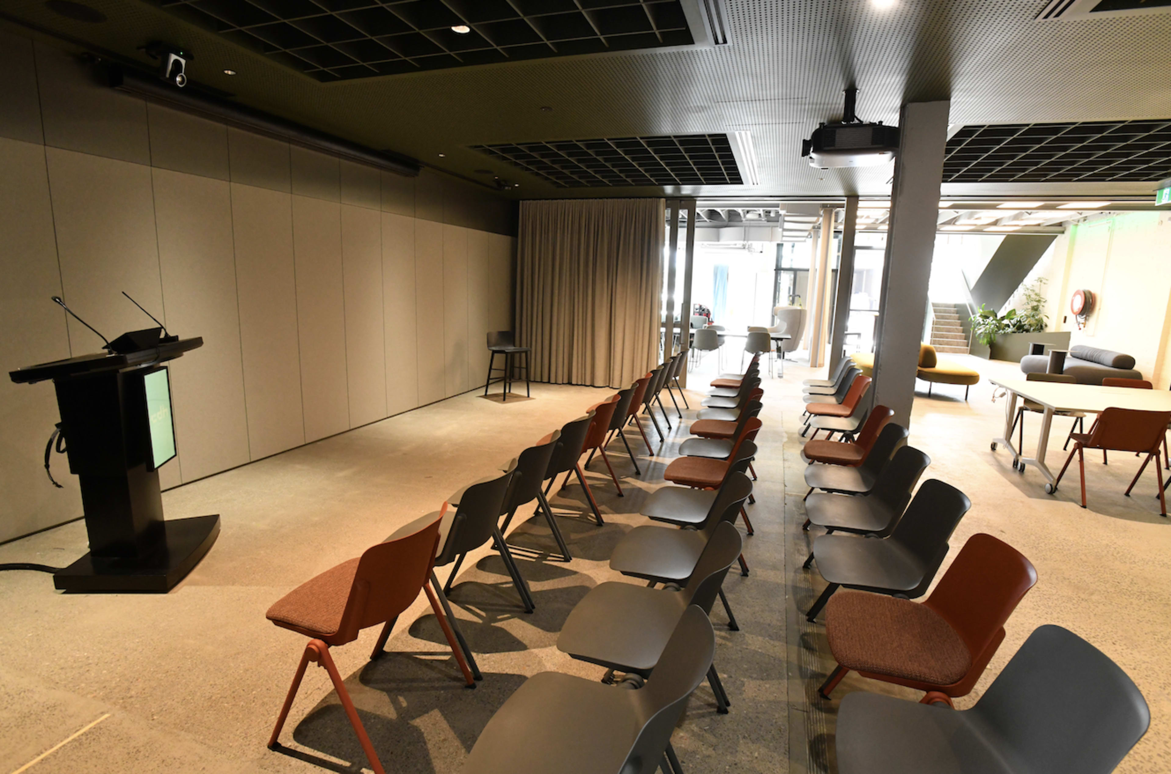 The image shows a conference room with rows of empty chairs facing a podium and a projector screen.
