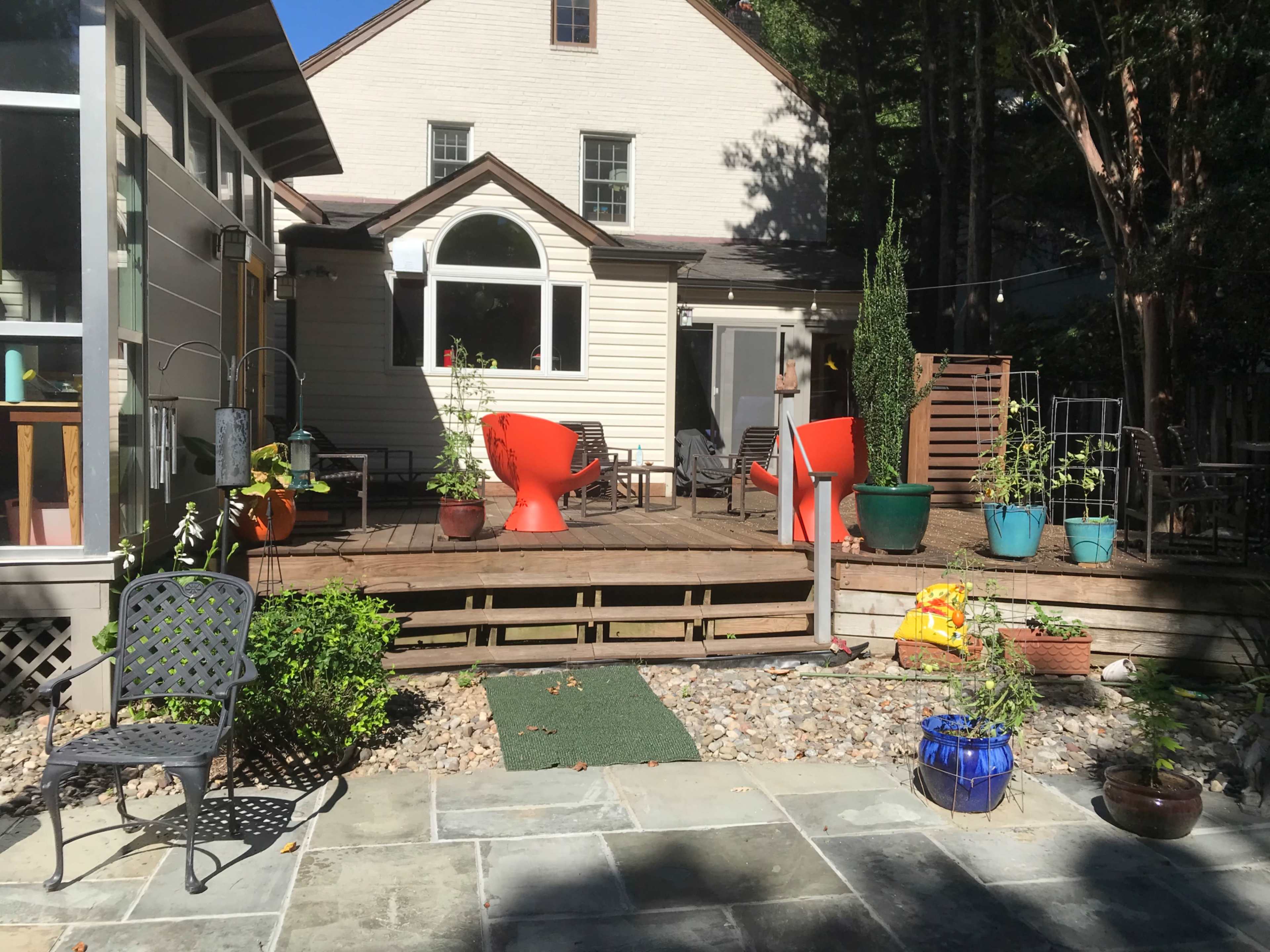 A backyard patio area with a wooden deck, two large red chairs, potted plants, and a stone pathway.