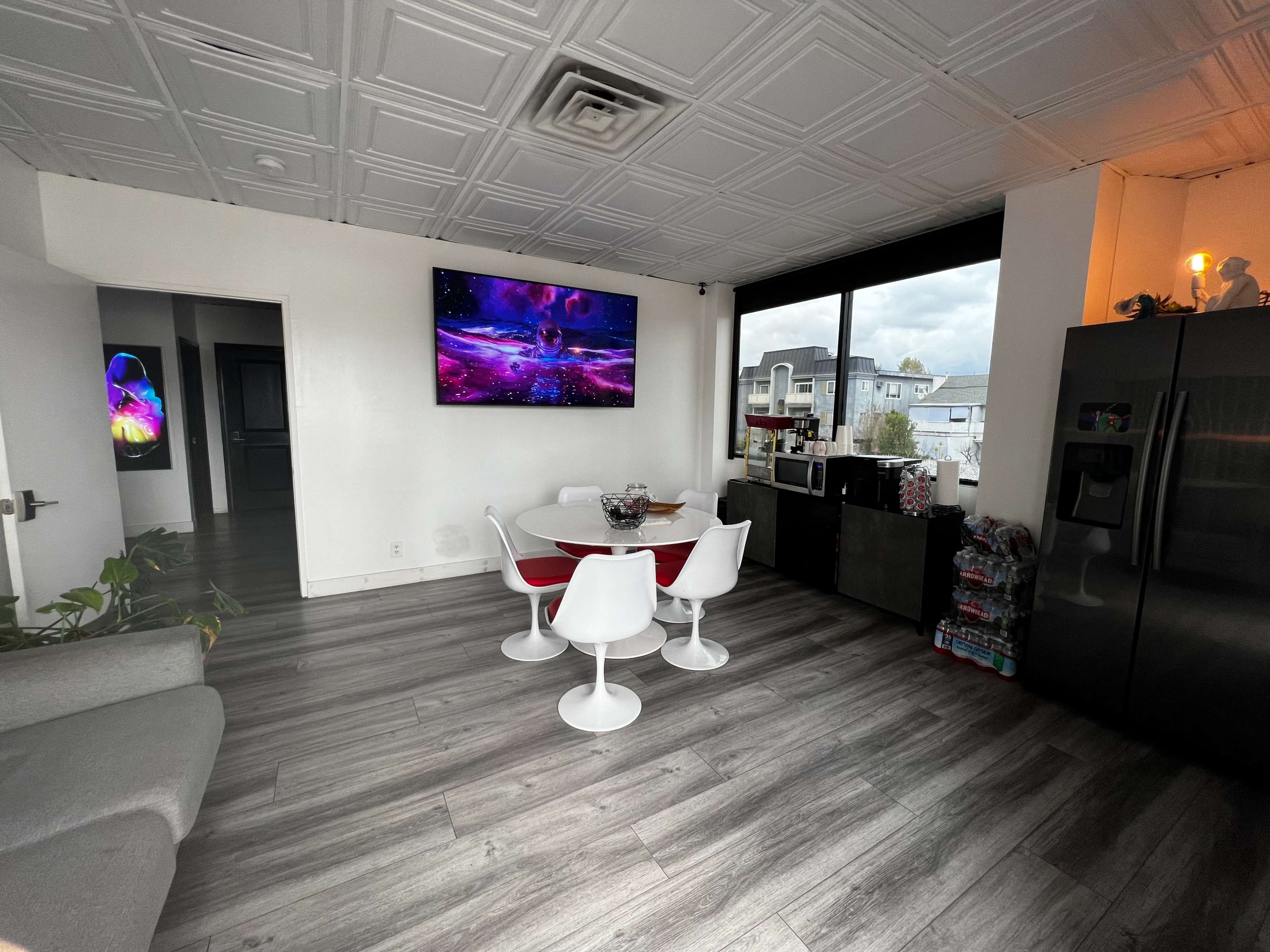 A modern kitchen and dining area features a round table with white chairs, a large wall-mounted television, and a refrigerator, all set against a backdrop of gray flooring and a window with an outdoor view.
