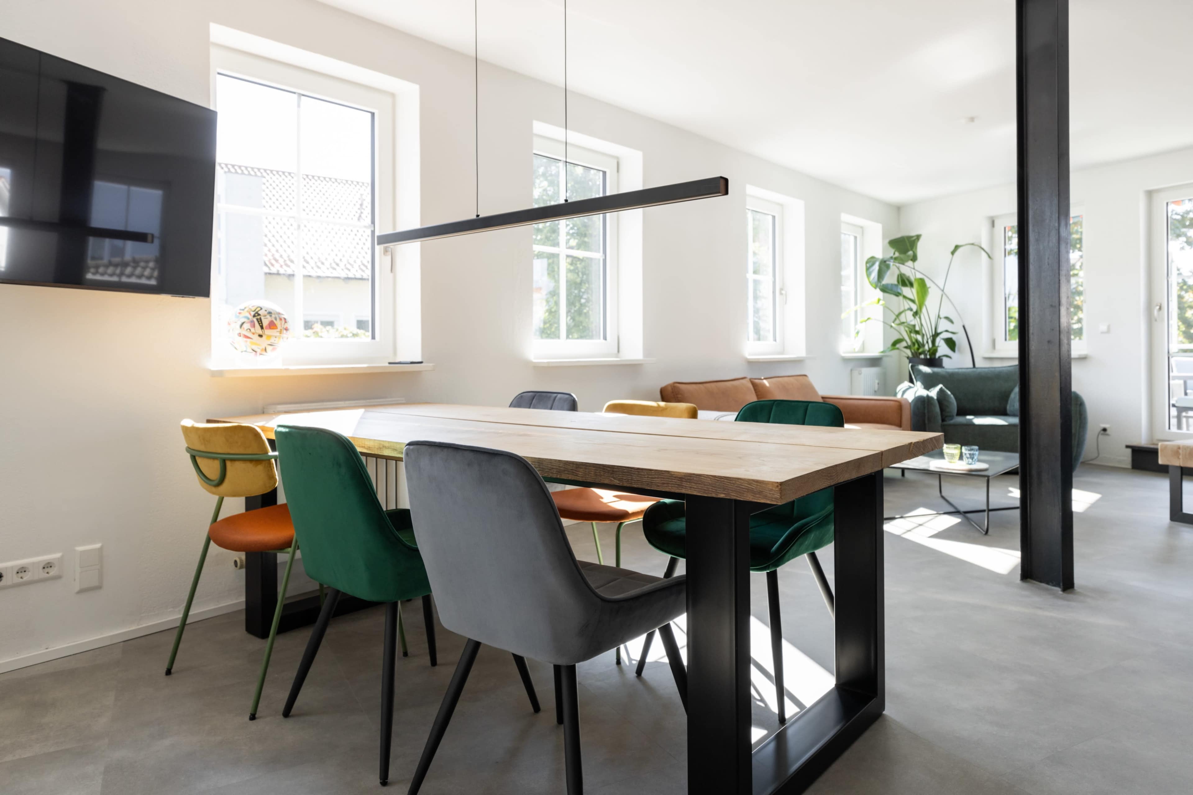 A modern dining area features a large wooden table surrounded by colorful chairs, with windows allowing natural light to fill the space.