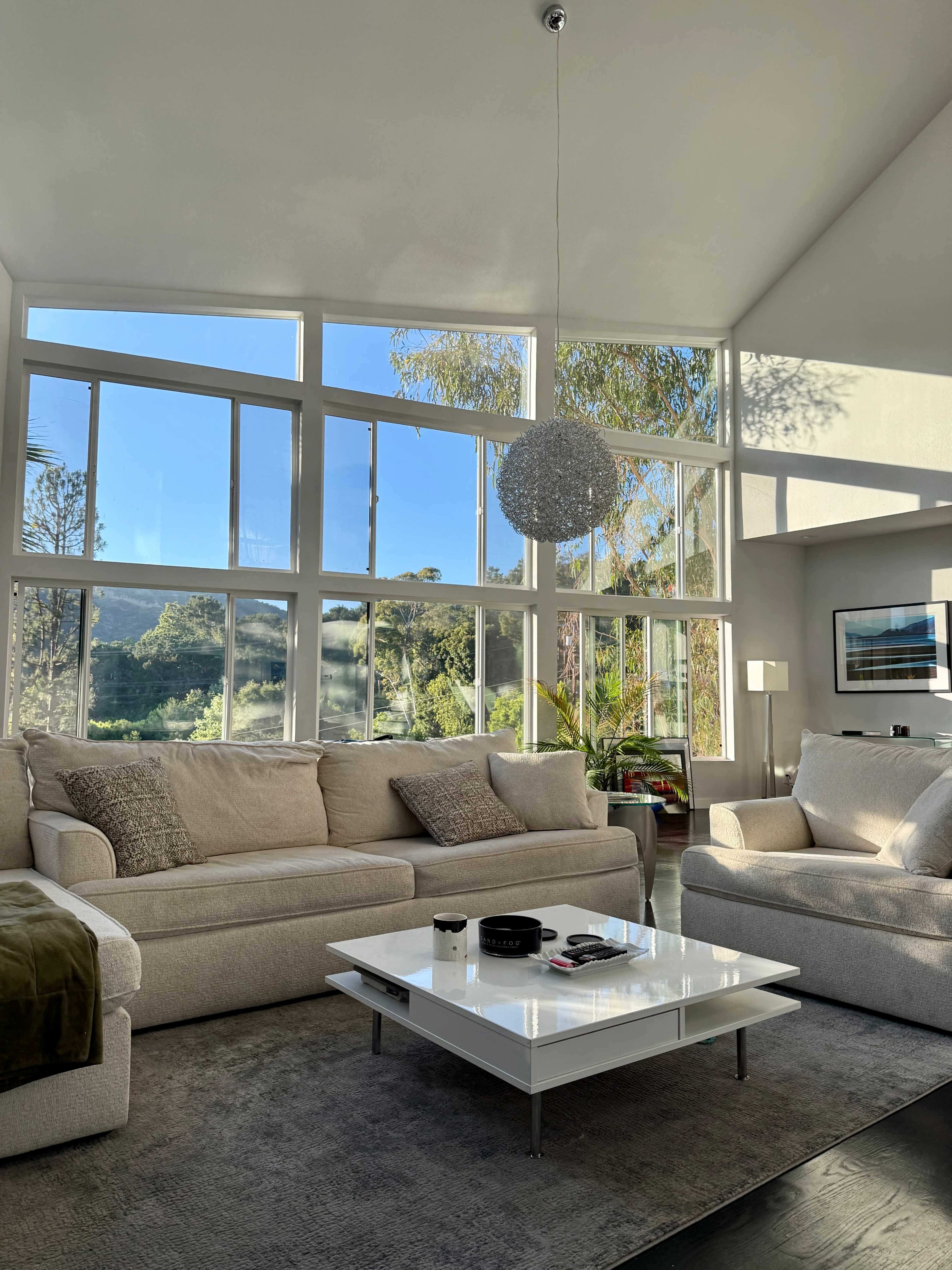 The living room features large windows that let in natural light, with a light-colored sofa set, a coffee table, and a textured area rug.