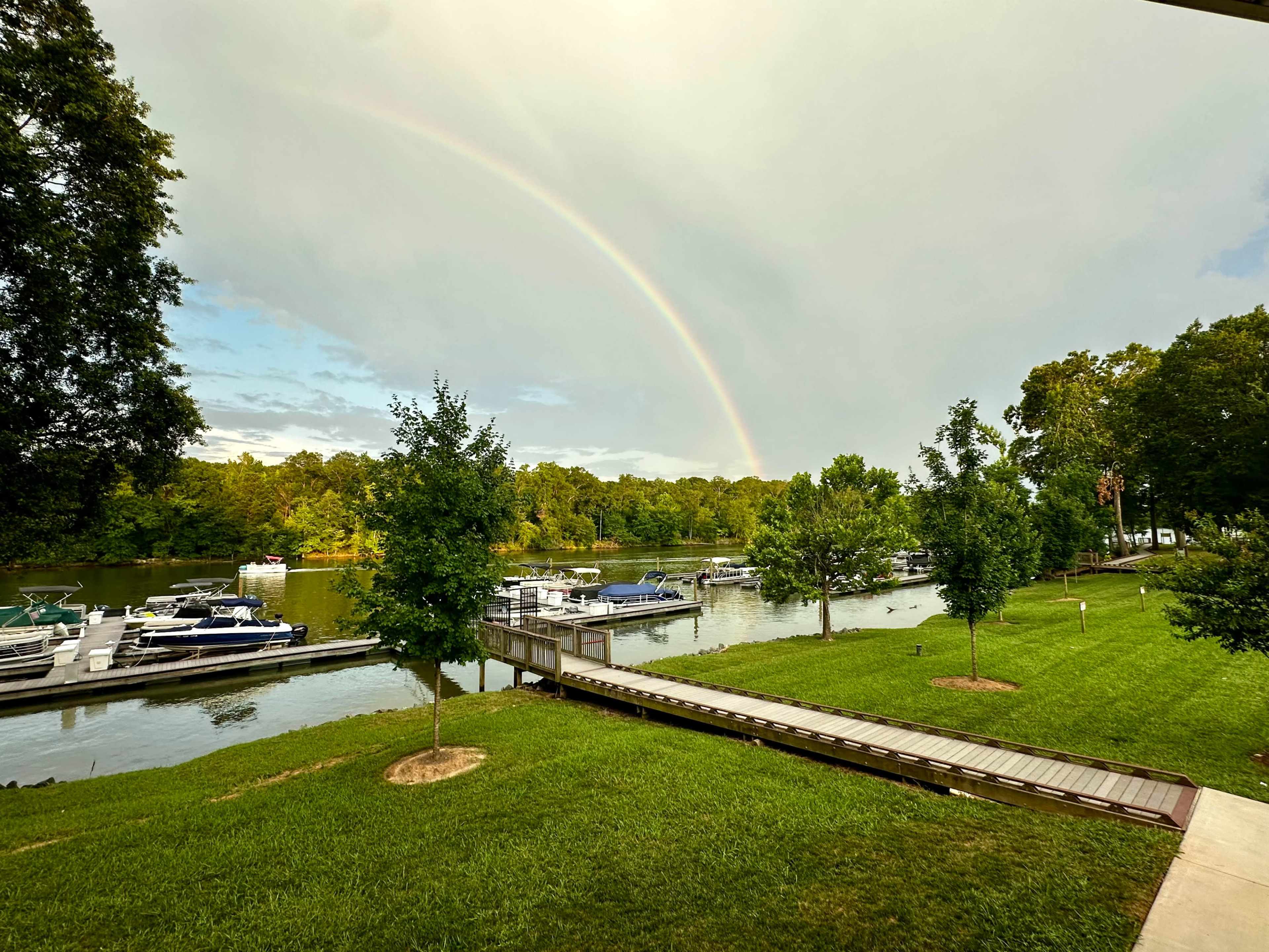 A rainbow arcs over a marina filled with boats, framed by trees and grass alongside a calm body of water.