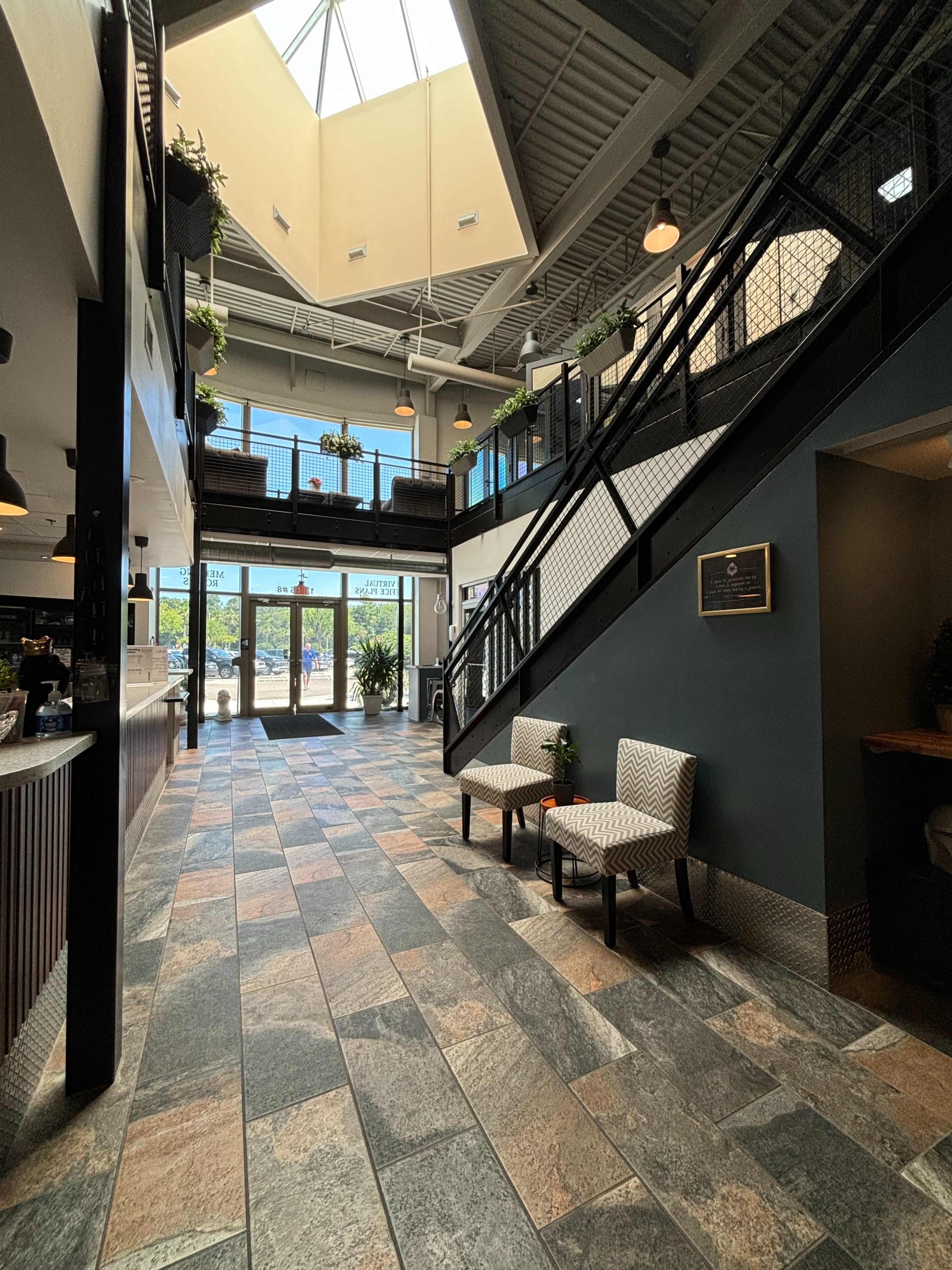 The spacious lobby features a two-story design with a skylight, a tiled floor, and a staircase leading to an upper level, complemented by potted plants and seating areas.