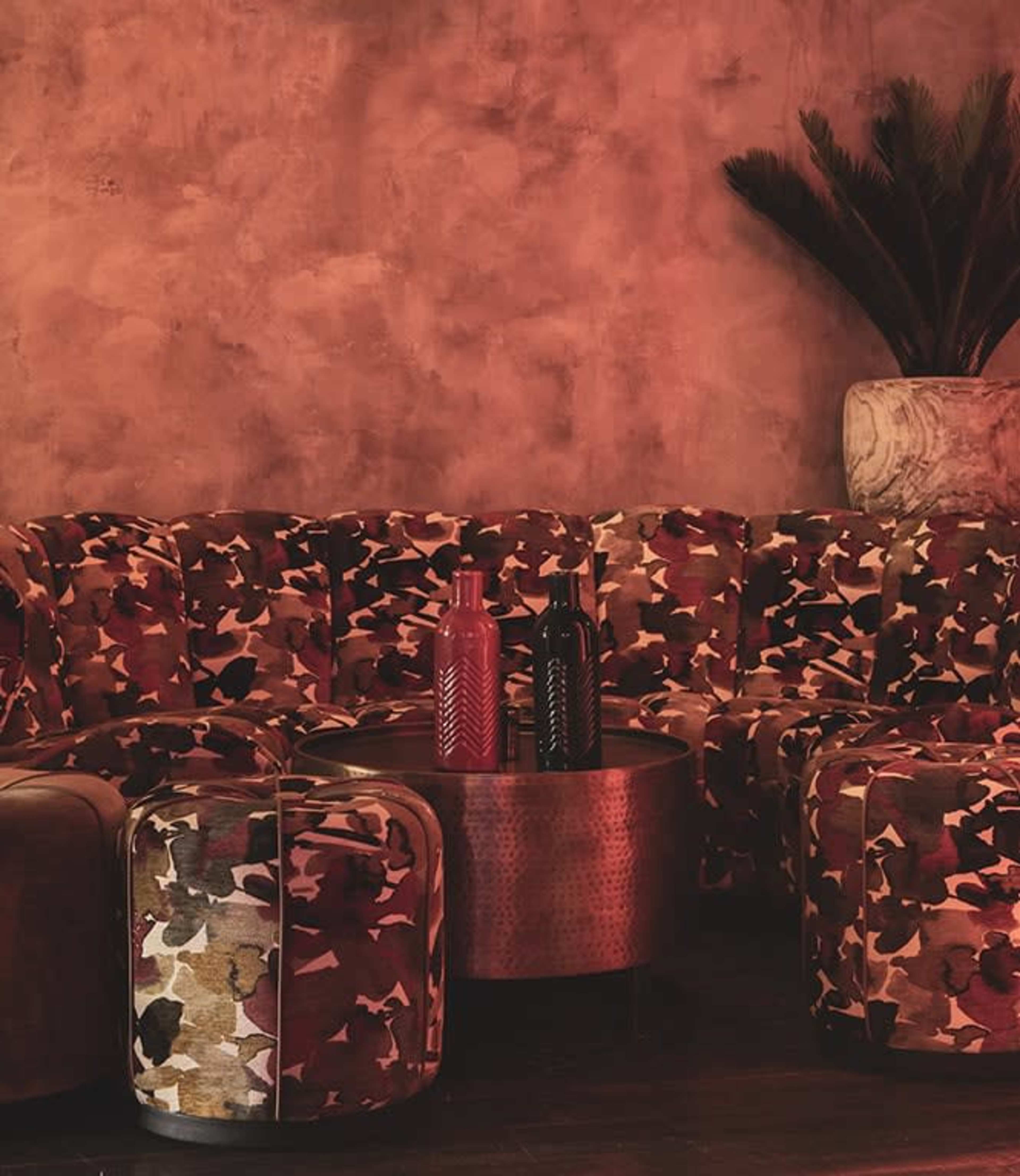 The image features a patterned sofa in earthy tones alongside a round, metallic table and a potted plant against a textured wall.