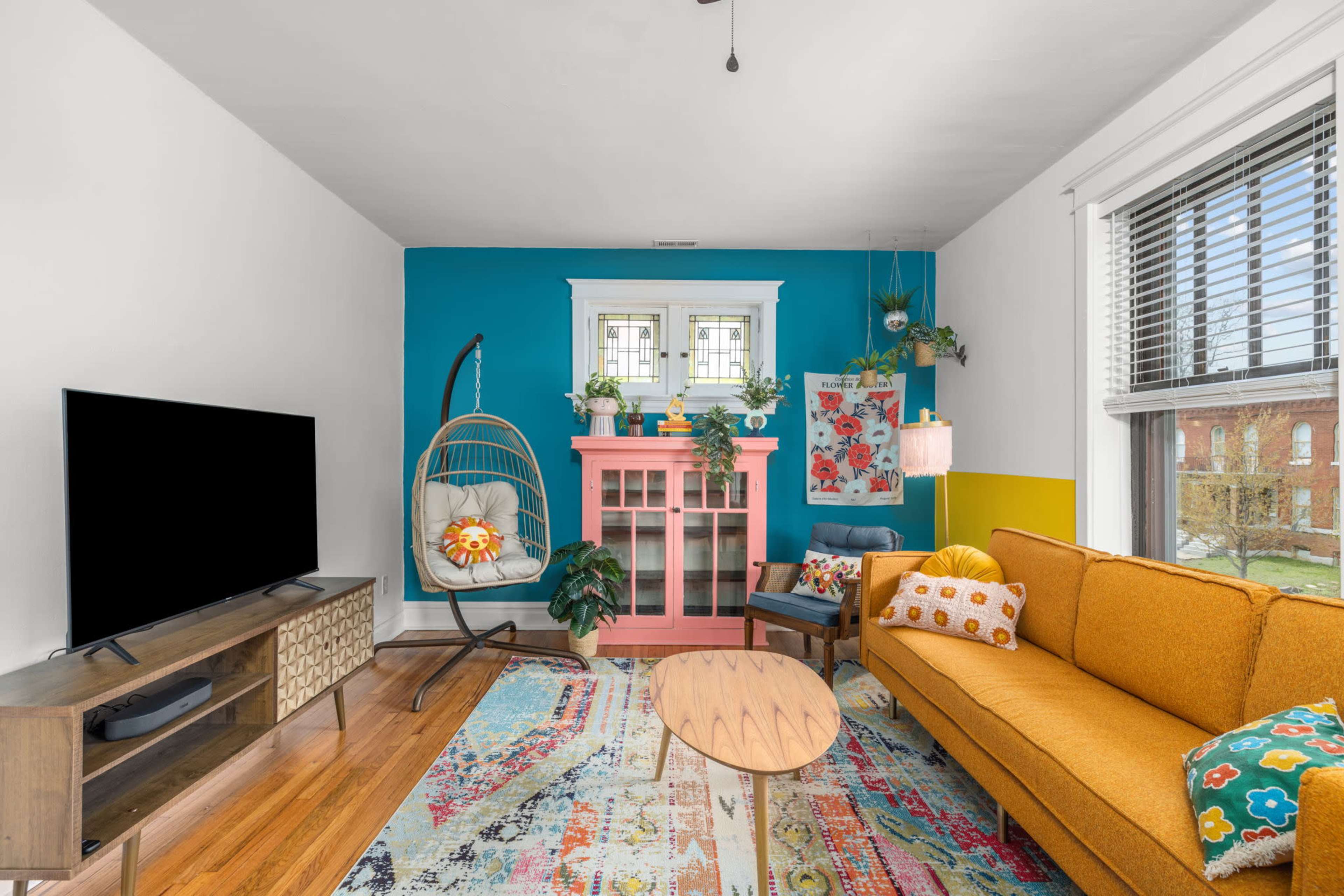 The living room features an orange sofa, a pink cabinet, a hanging chair, and colorful decor against a blue accent wall.