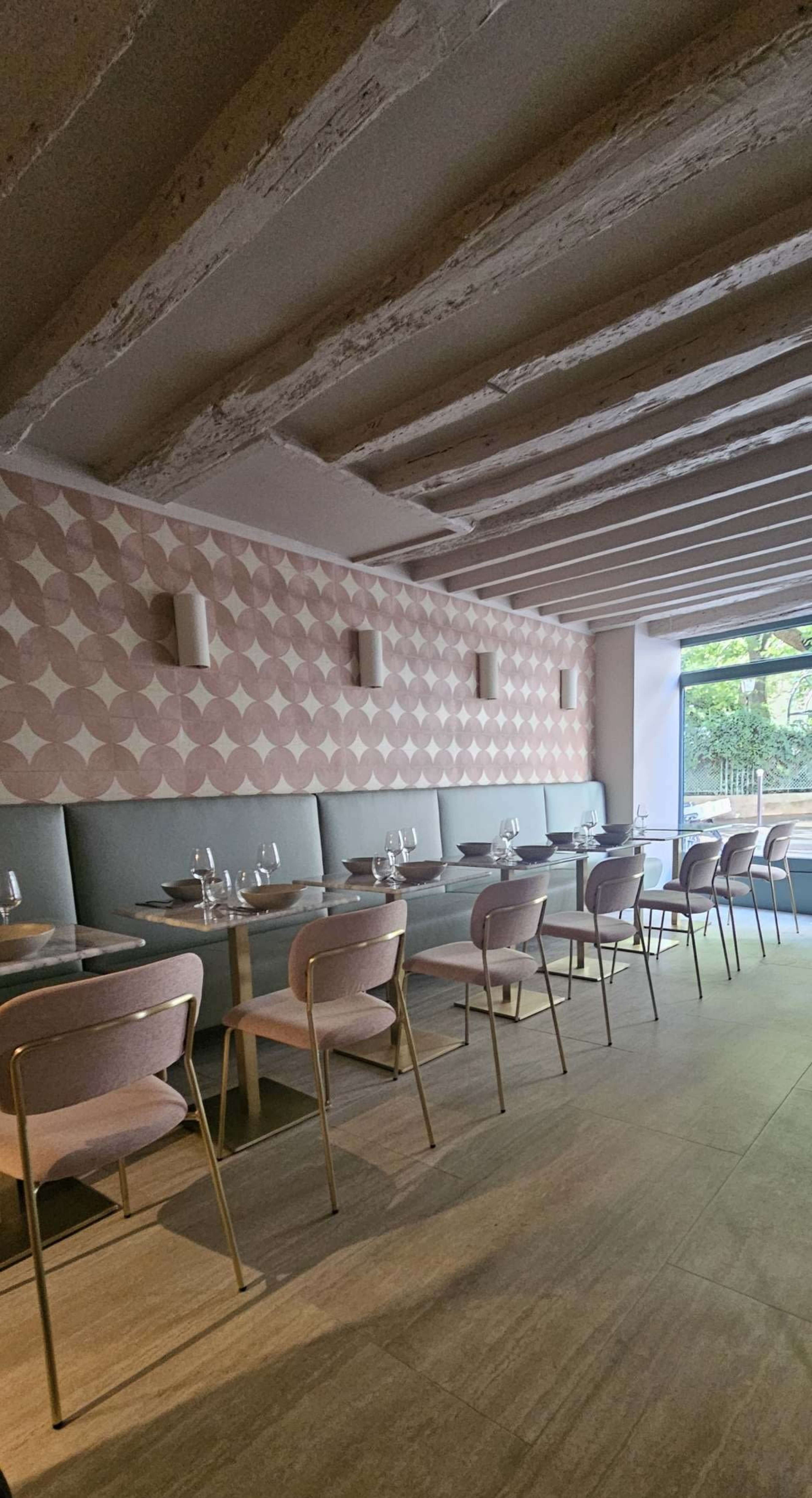 The image shows a modern restaurant interior featuring a patterned pink wall, gray seating, and wooden tables set with glassware.