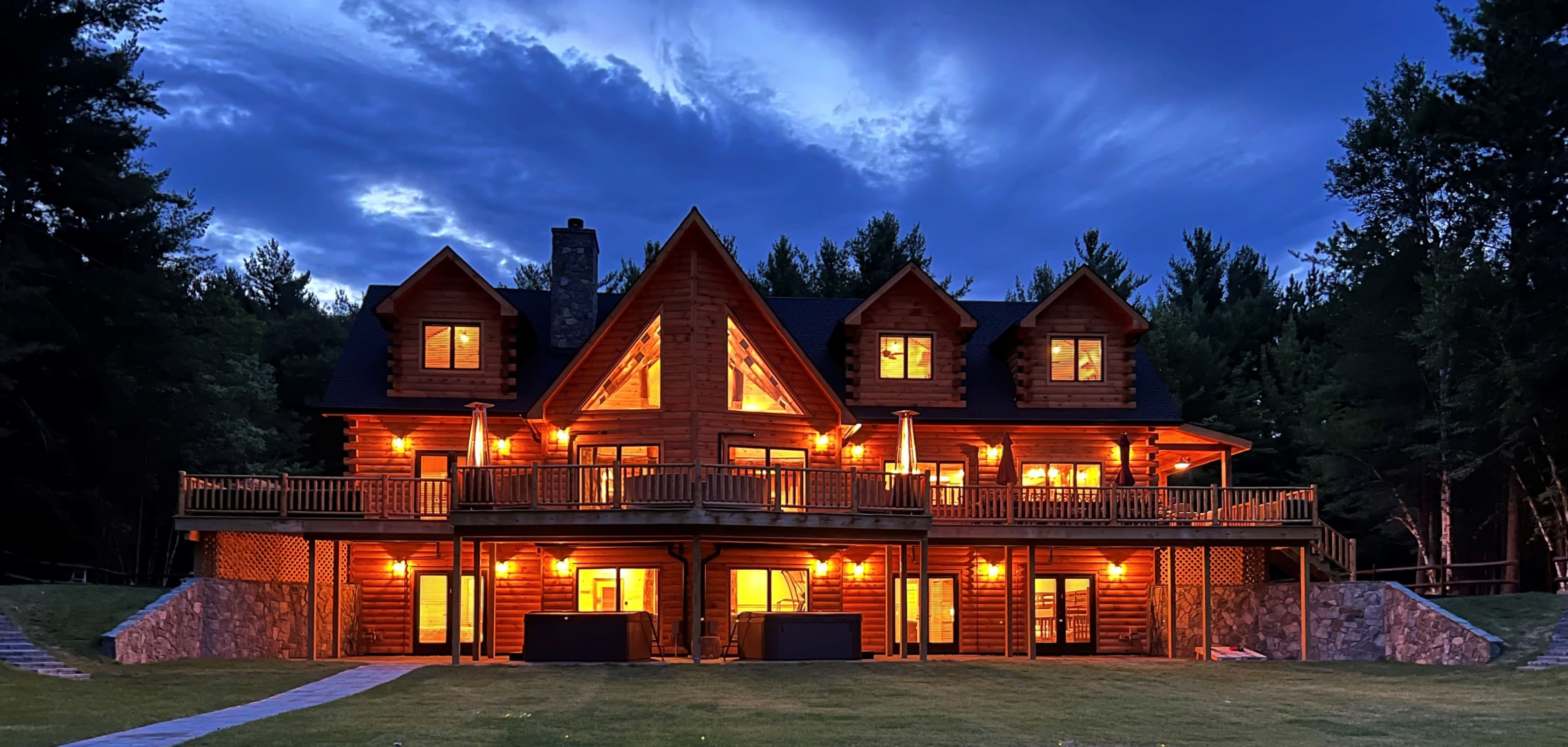 A large wooden cabin with multiple gables, illuminated by warm lights, set against a twilight sky filled with dramatic clouds.
