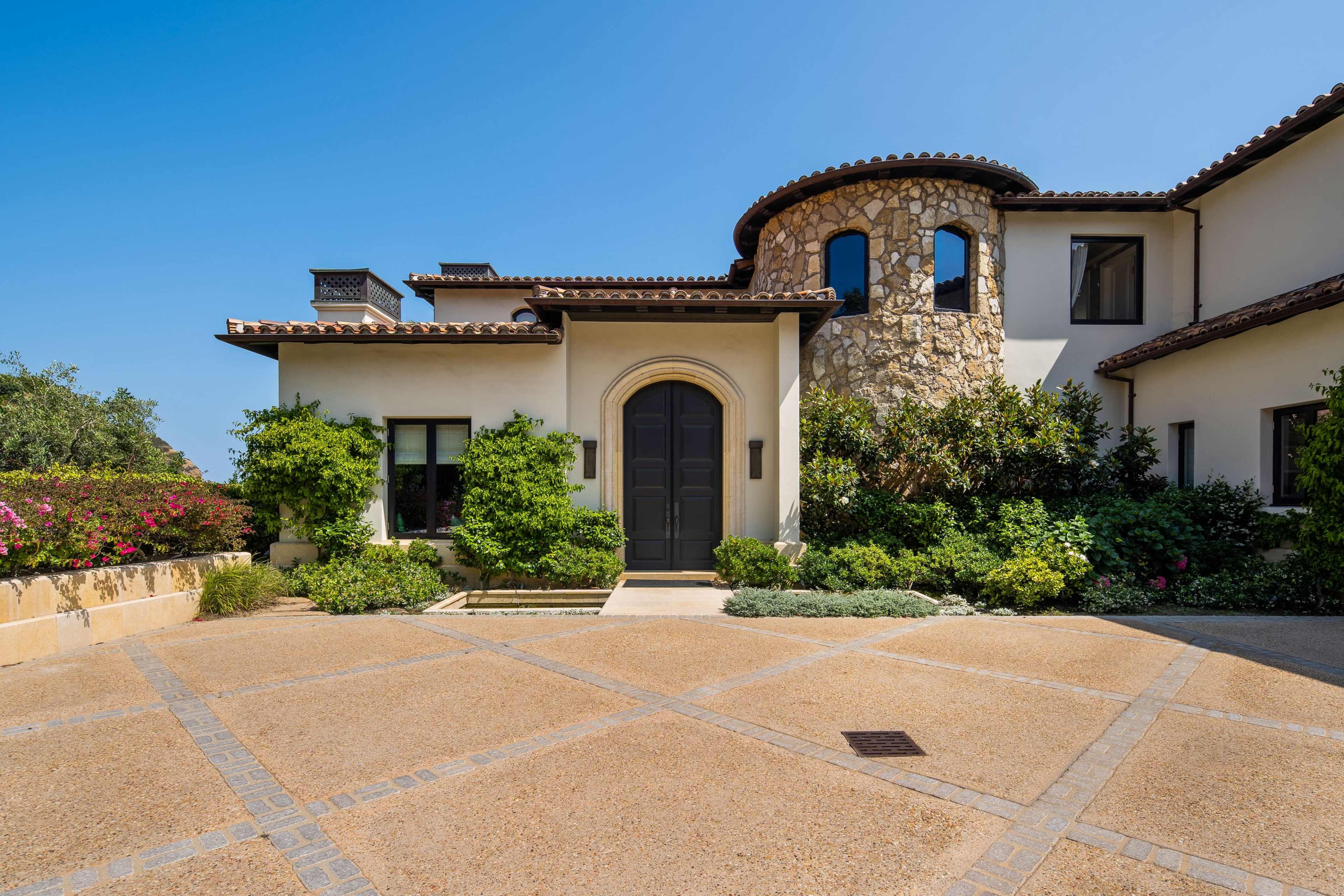 The image shows a stone and stucco house with a circular tower, surrounded by landscaped gardens and a paved driveway.