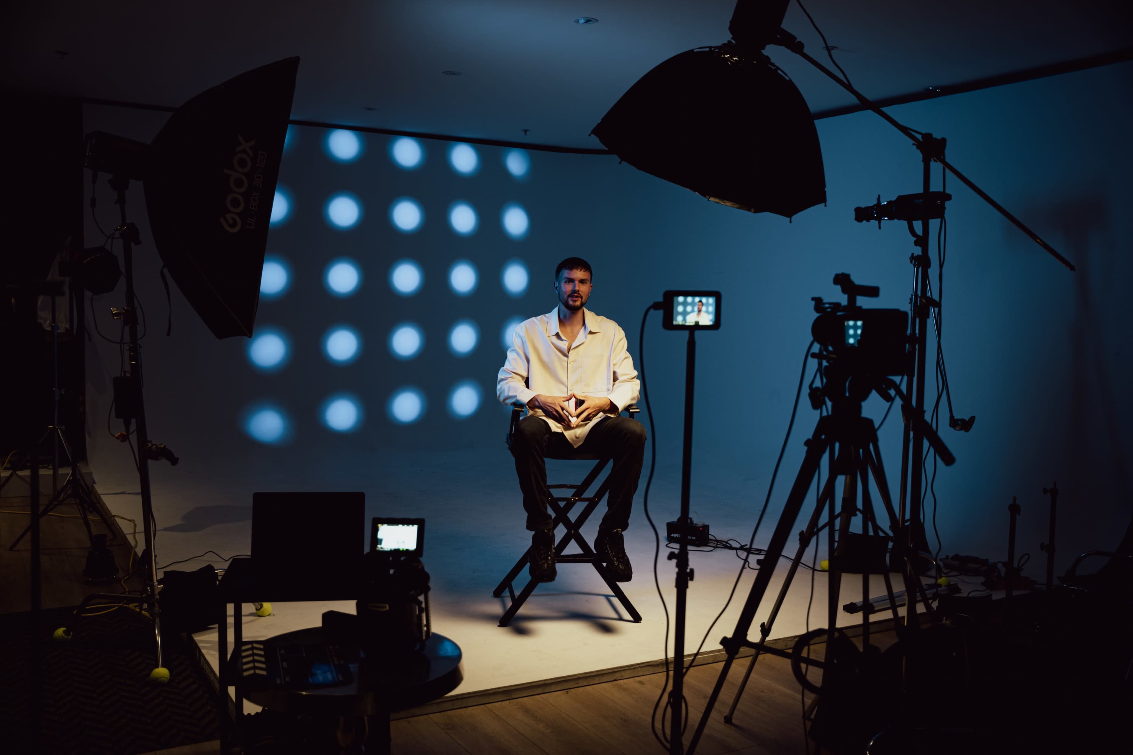 A man sits on a director's chair in a well-lit studio, surrounded by various camera equipment and lighting setups.