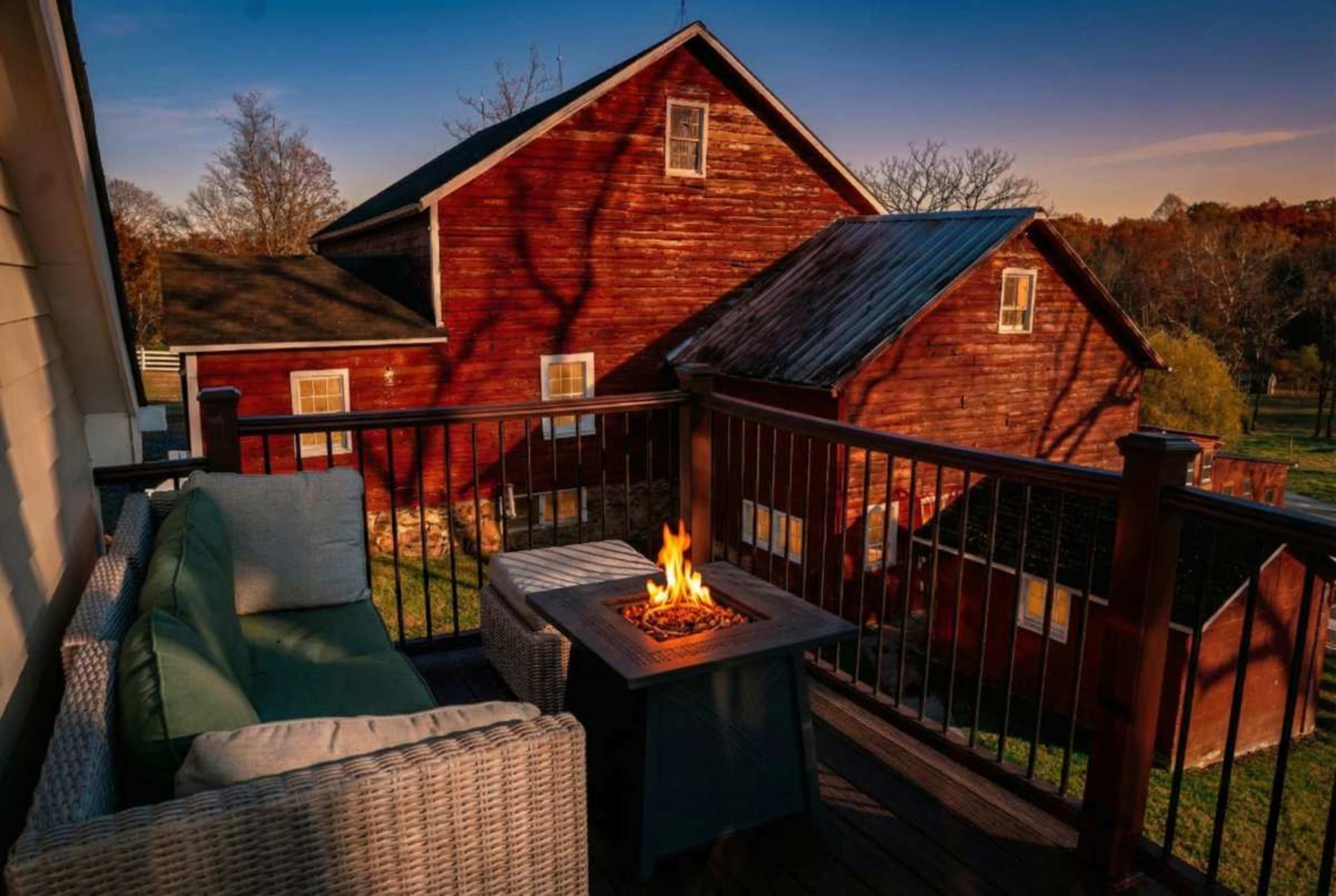 A cozy outdoor seating area with a fire pit overlooks a rustic red barn at sunset.