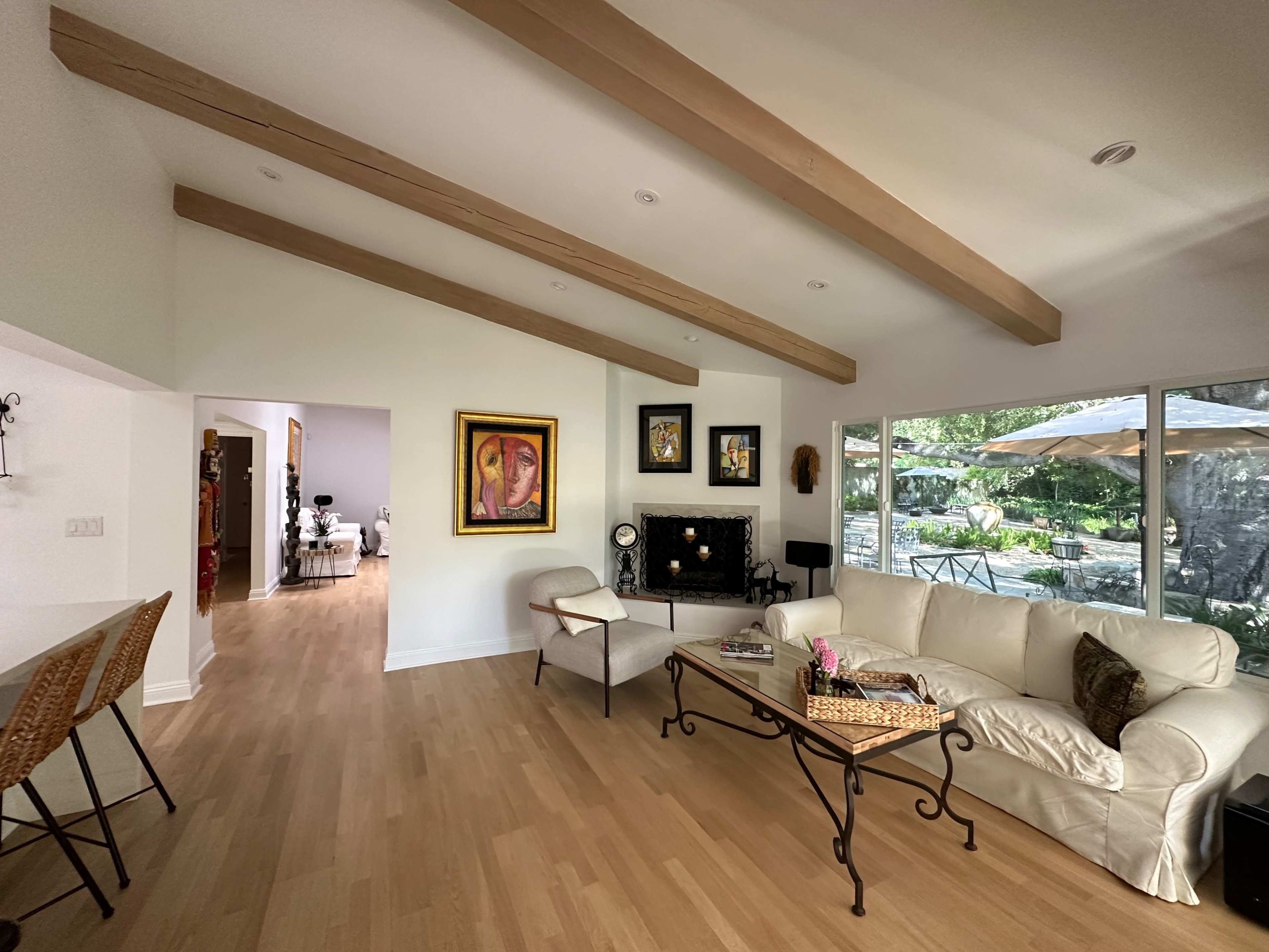A spacious living room features a white couch, a coffee table, and wooden beams on the ceiling, with large windows overlooking an outdoor patio area.